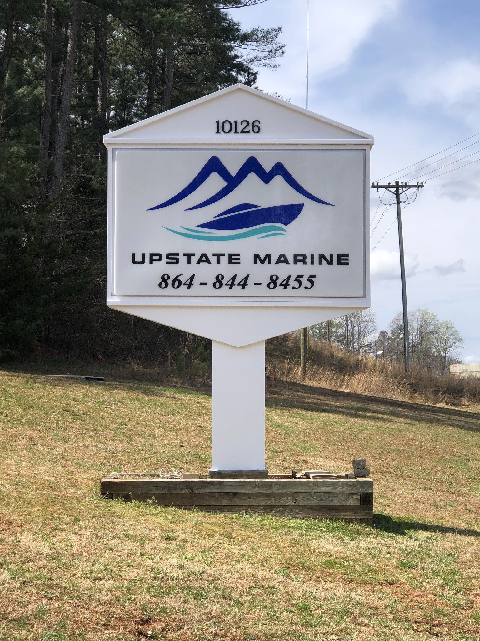 A large white sign with a blue mountain and boat logo, displaying the text "UPSTATE MARINE" and a phone number, located outdoors on a grassy area with trees and power lines in the background.
