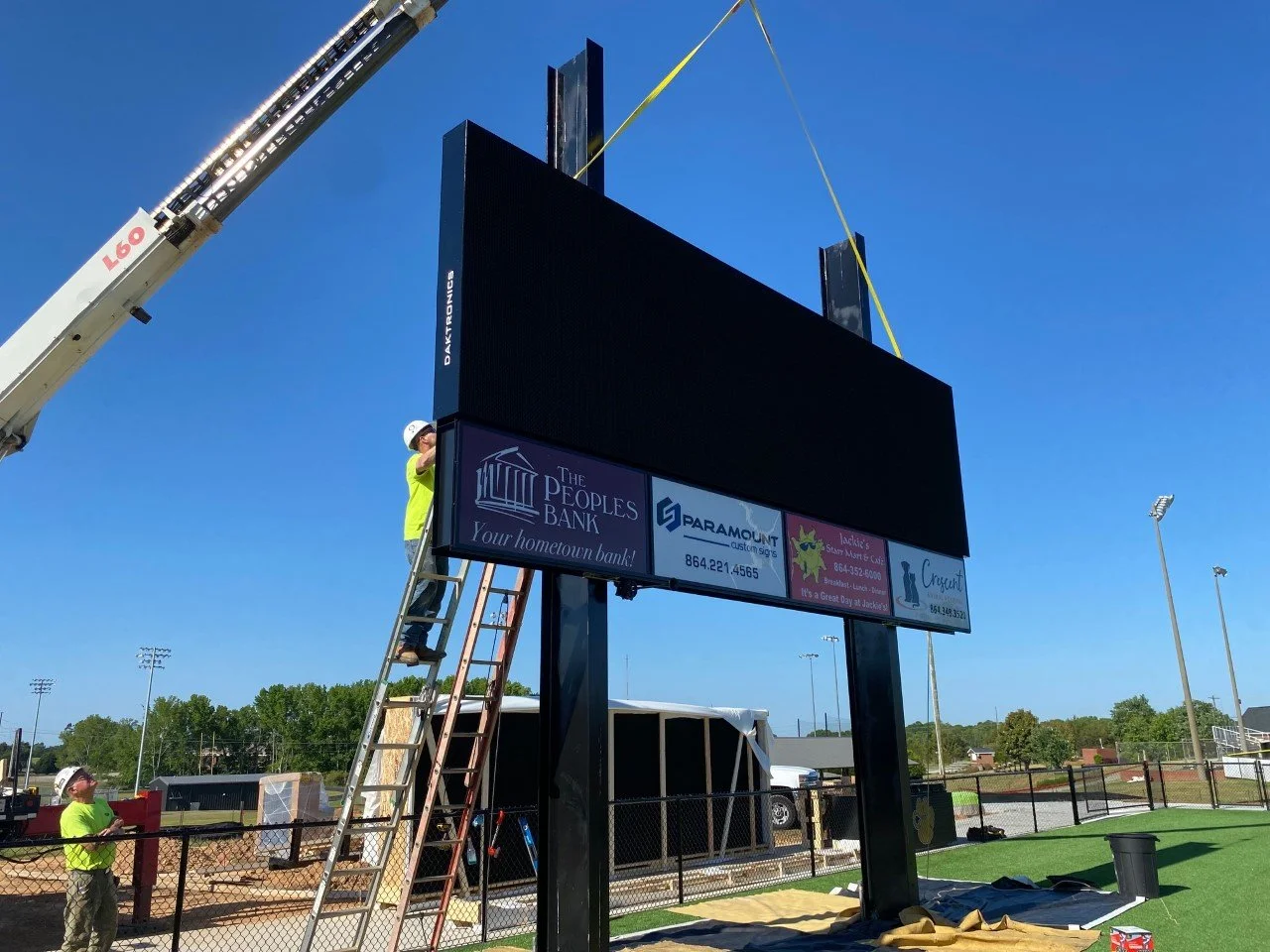 Construction workers installing a large outdoor electronic scoreboard on a sports field under a clear blue sky.