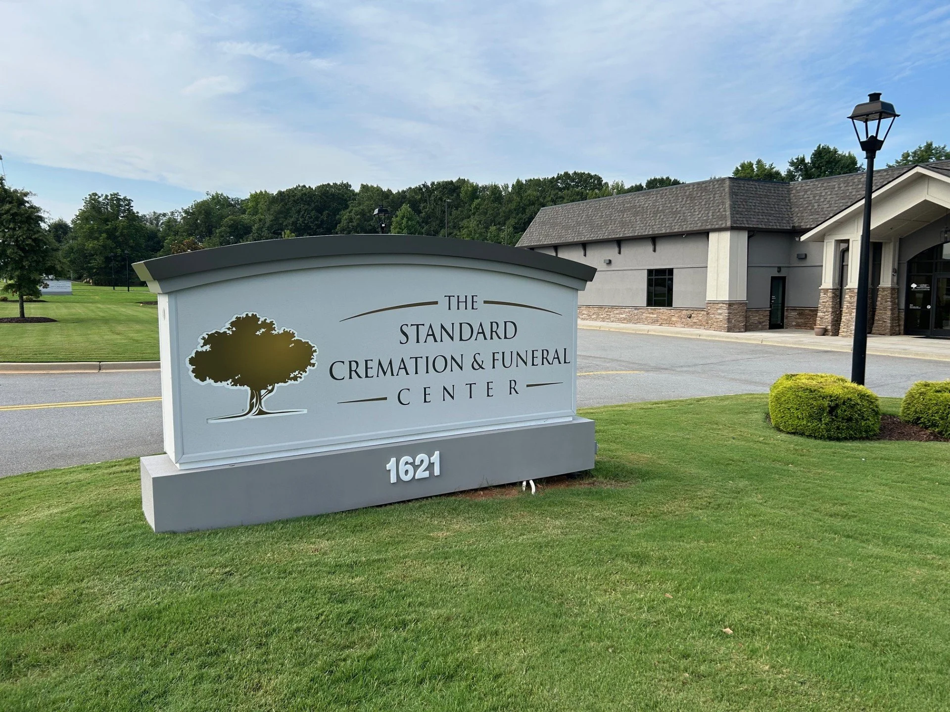 Sign for The Standard Cremation & Funeral Center with a tree logo, located at 1621, in front of a building with gray walls, a shingled roof, and well-maintained lawn.