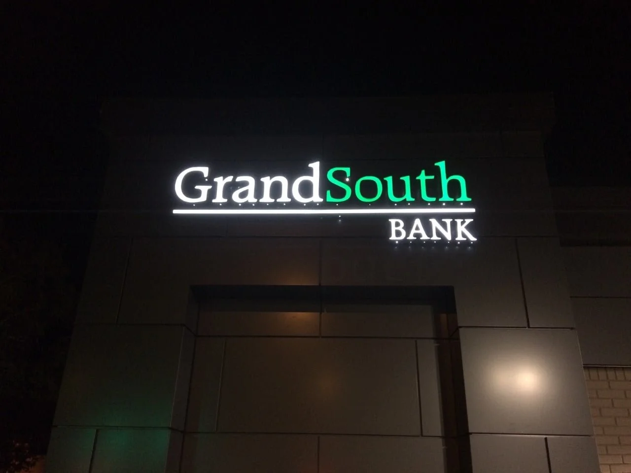 Night view of a sign for Grand South Bank with illuminated white and green letters on a dark building.