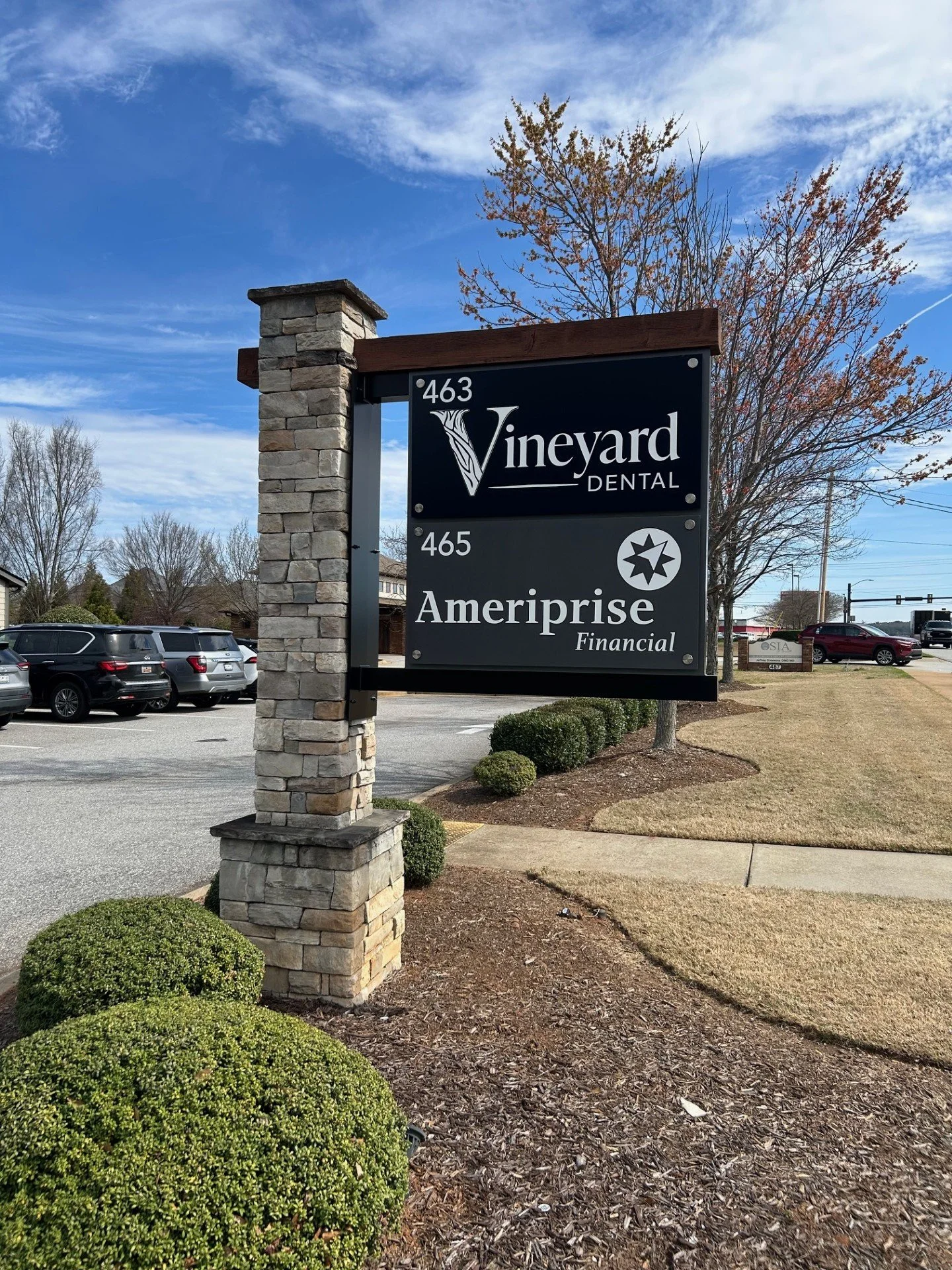 Signboard advertising Vineyard Dental and Ameriprise Financial at a roadside with parked cars and trees in the background.