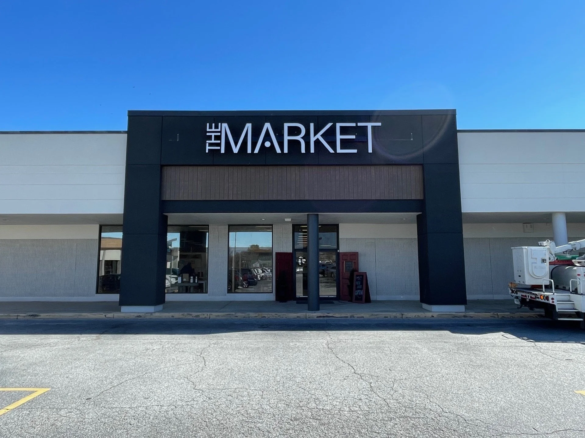 Front view of a grocery store named 'The Market' with a black sign and modern design, under a clear blue sky.