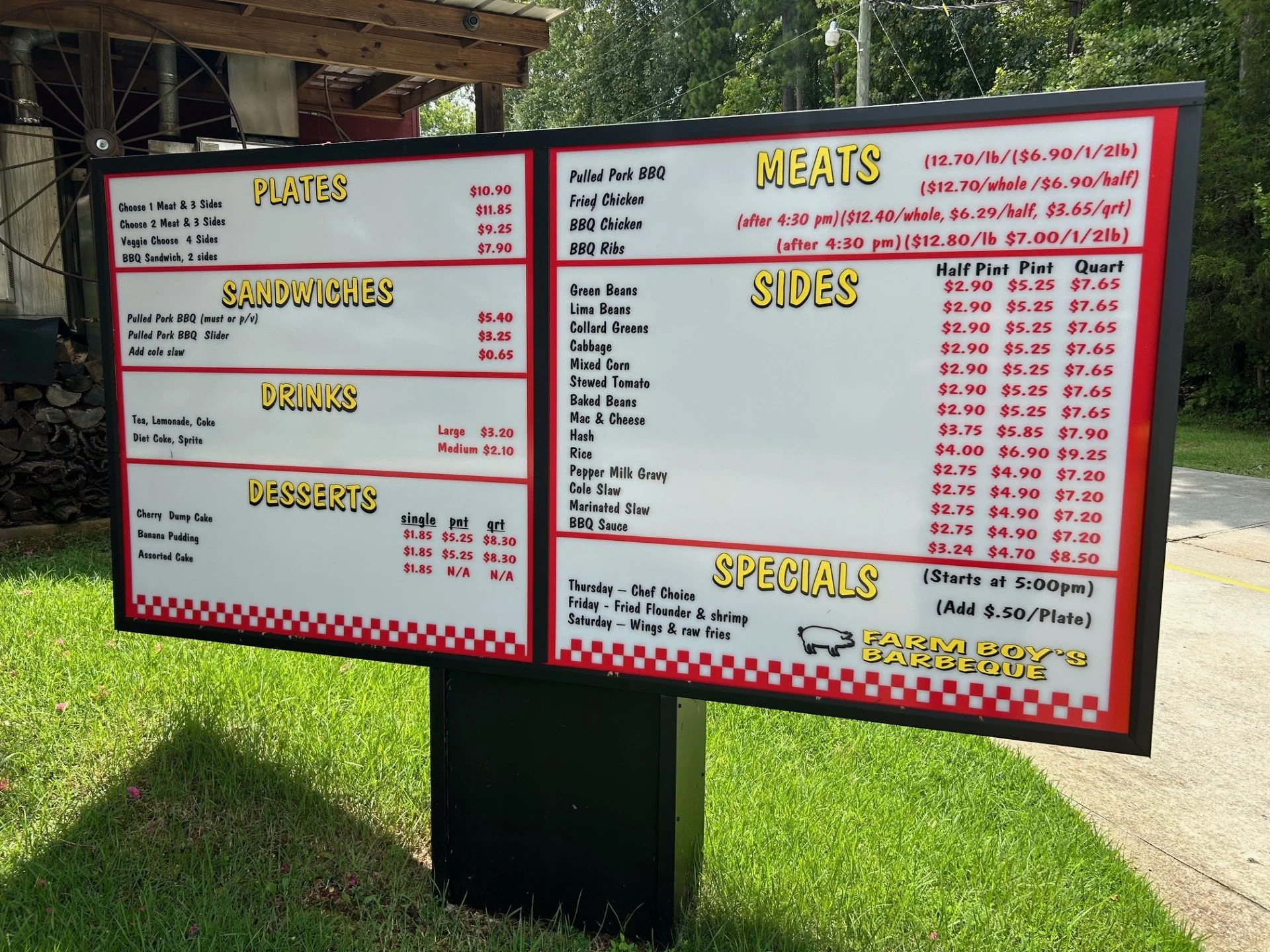 A black and white restaurant menu board outdoors displaying various barbecue plates, sandwiches, drinks, sides, desserts, and specials, with prices listed for different portion sizes and times.