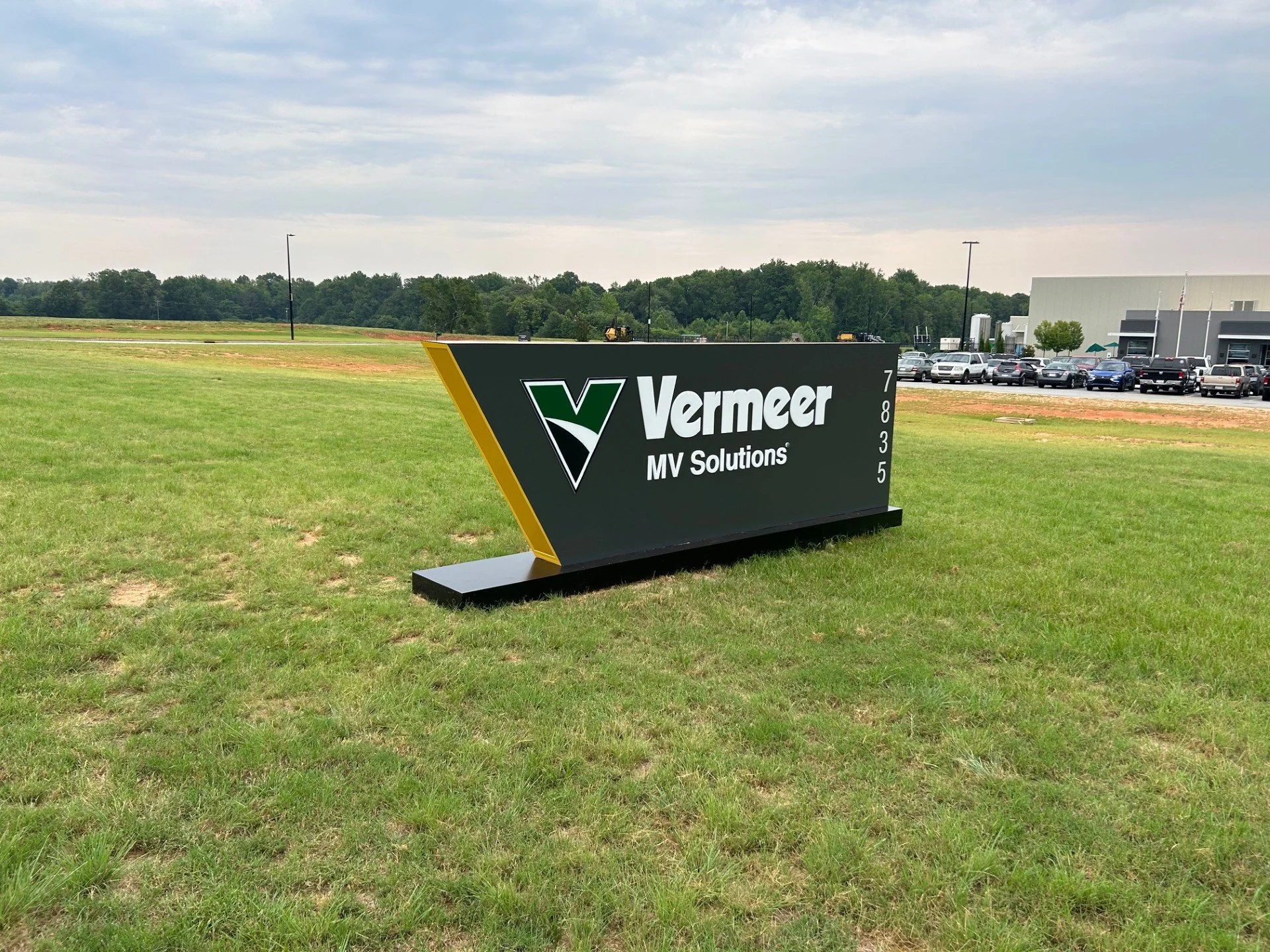 A black and green sign on a grassy field with the Vermeer logo and the words "Vermeer MV Solutions". In the background, there is a parking lot with cars, a building, and some trees under a cloudy sky.
