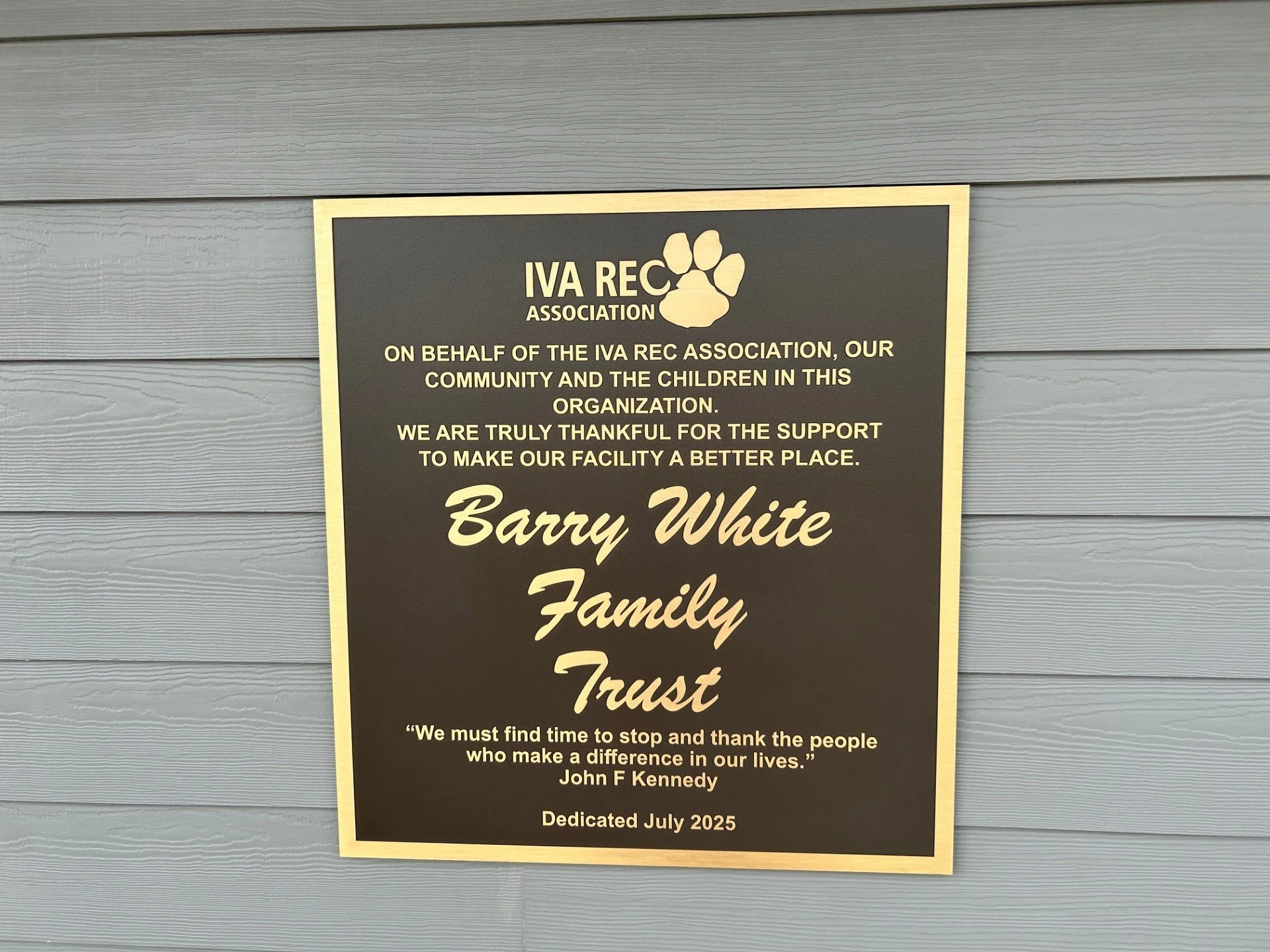 A memorial plaque on a gray wooden wall honoring the Barry White Family, with a quote by John F. Kennedy and a dedication date of July 2025.