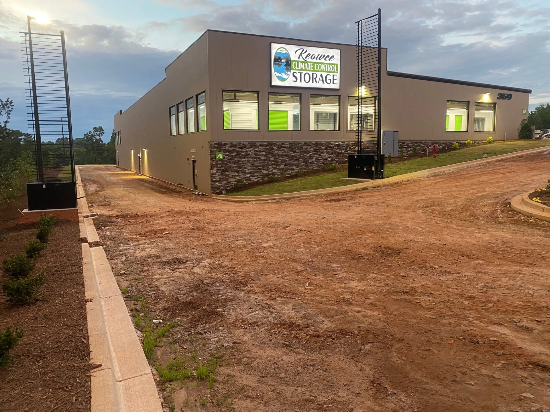 A modern building with a sign that reads 'Keowee Climate Control Storage'. The building has large windows and a stone accent on the lower part of the exterior wall. The surrounding area is under development with a dirt road and a landscaped strip wit