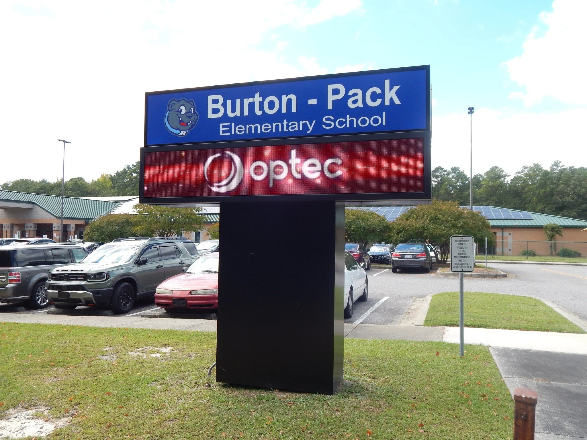 A parking lot with multiple cars parked and a school sign that reads 'Burton-Pack Elementary School' with a bear mascot. There is an electronic sign below it displaying the word 'optec' with a red background. Trees and a building with solar panels ca