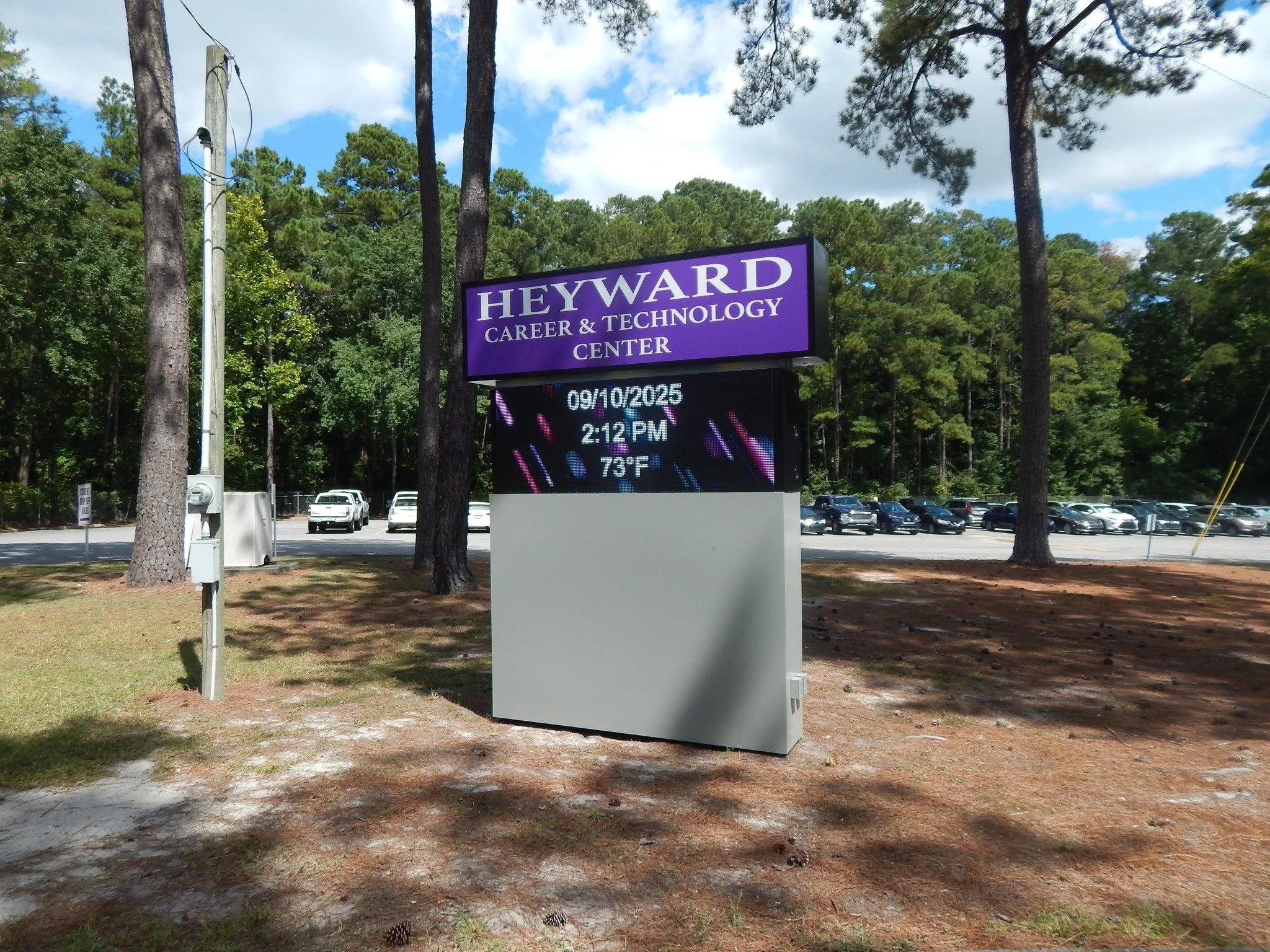 Outdoor parking lot surrounded by tall pine trees with a digital signboard displaying the date, time, and weather temperature, and a purple college sign that reads "HEYWARD CAREER & TECHNOLOGY CENTER."