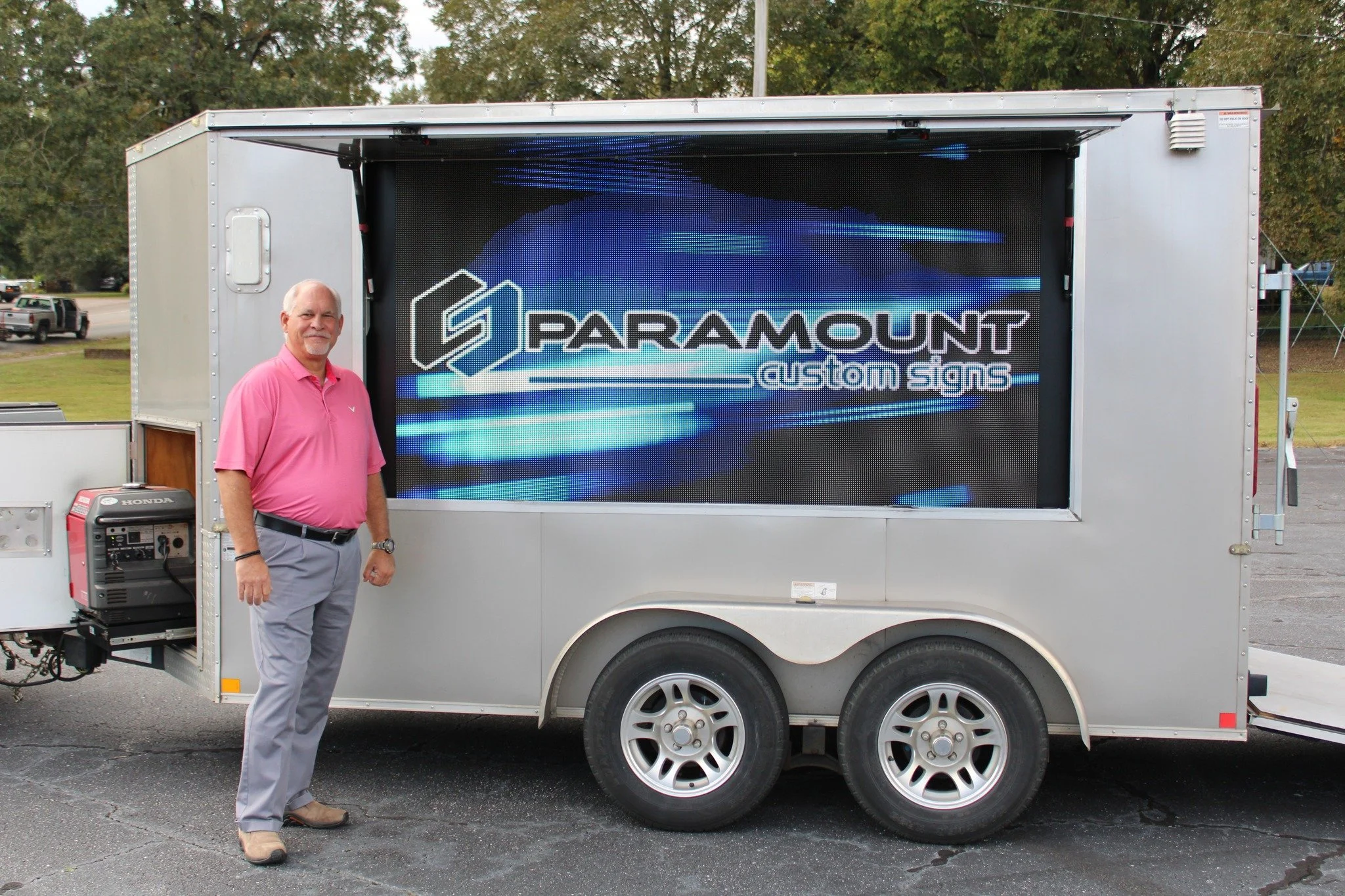 A man standing next to a mobile digital sign with the logo and name 'Paramount Custom Signs' displayed on it, outdoors in a parking lot with trees in the background.