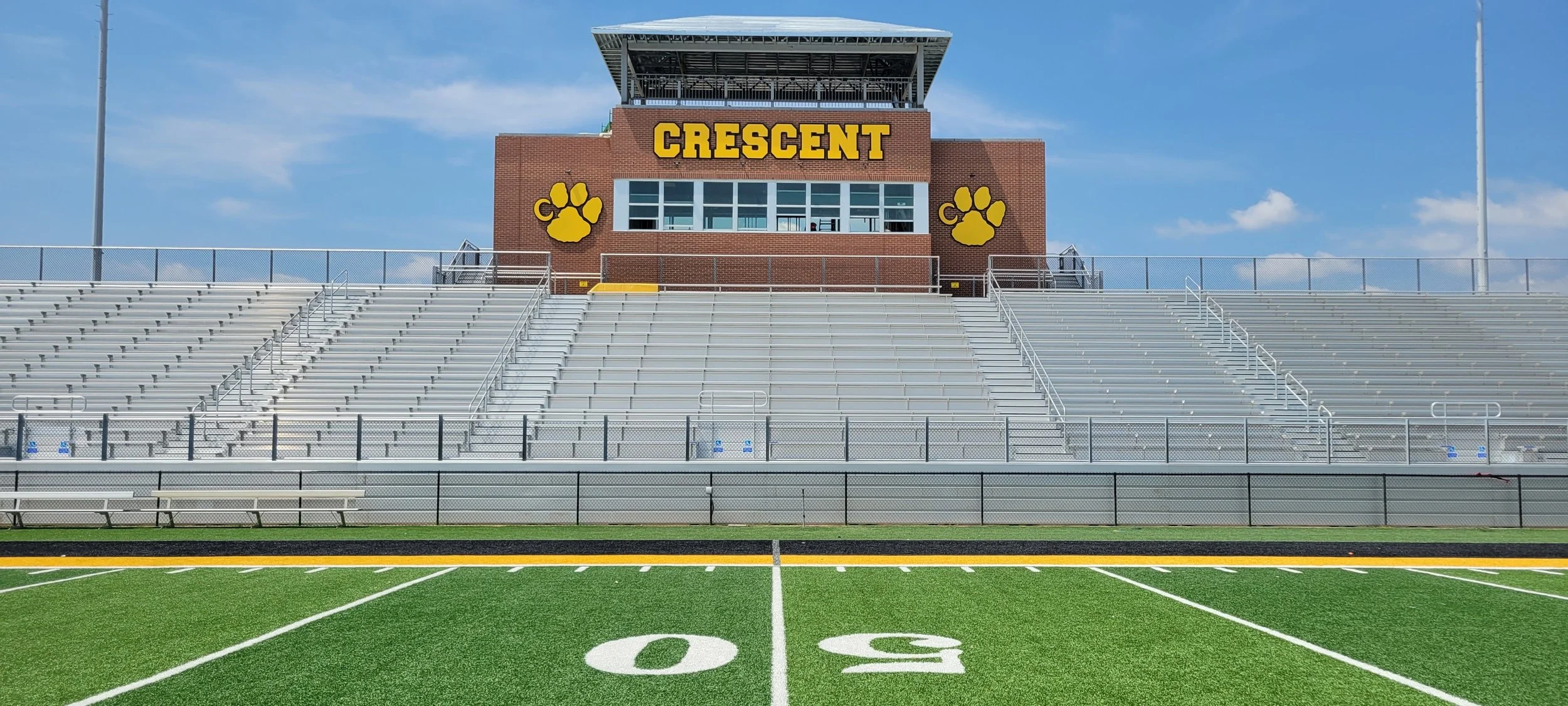 Empty football stadium with bleachers and a red brick press box labeled 'Crescent' with bear paw prints, under a blue sky.