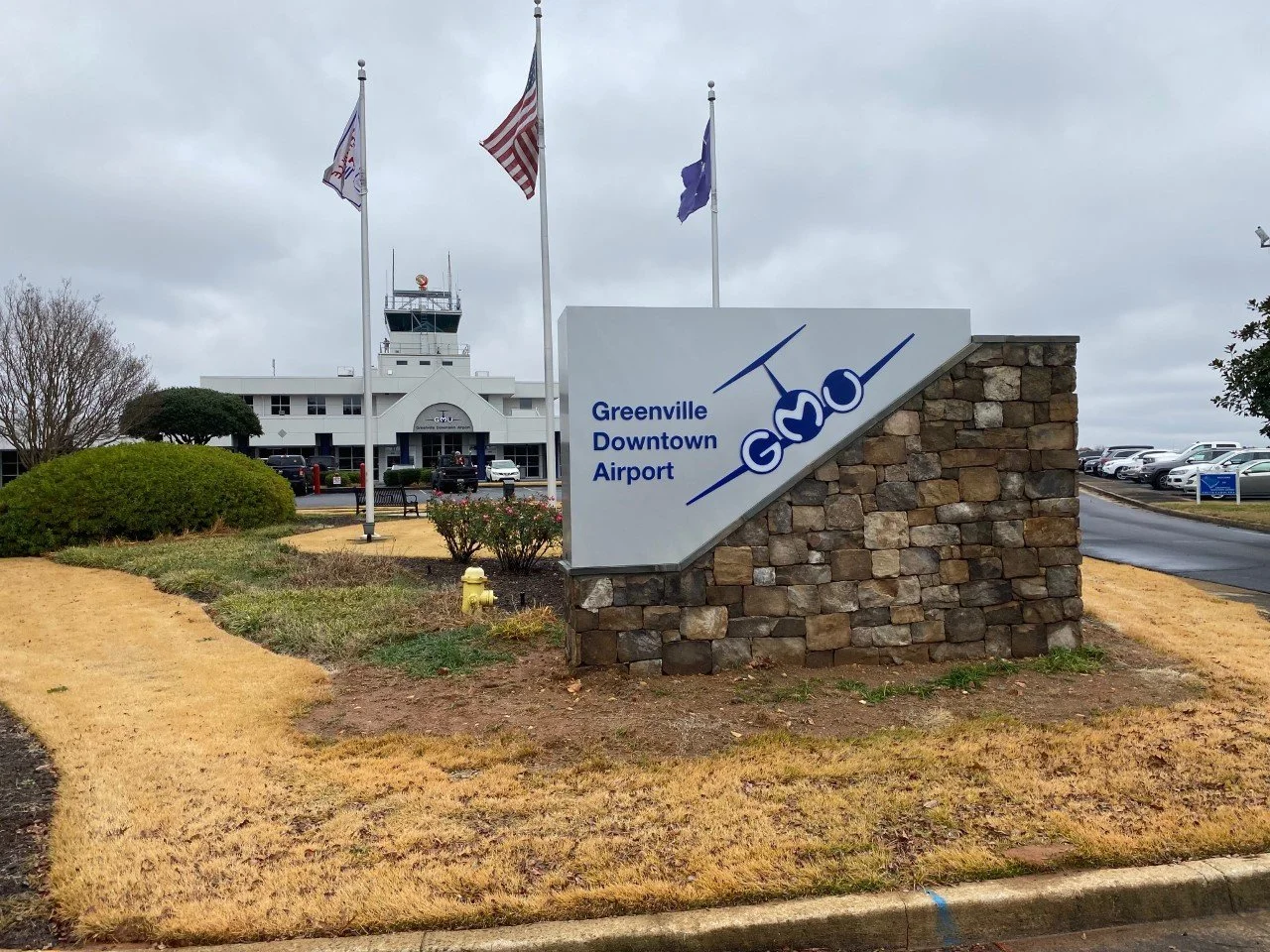 Sign for Greenville Downtown Airport with the words and airplane graphic, with the airport terminal building and parking lot in the background under cloudy sky.