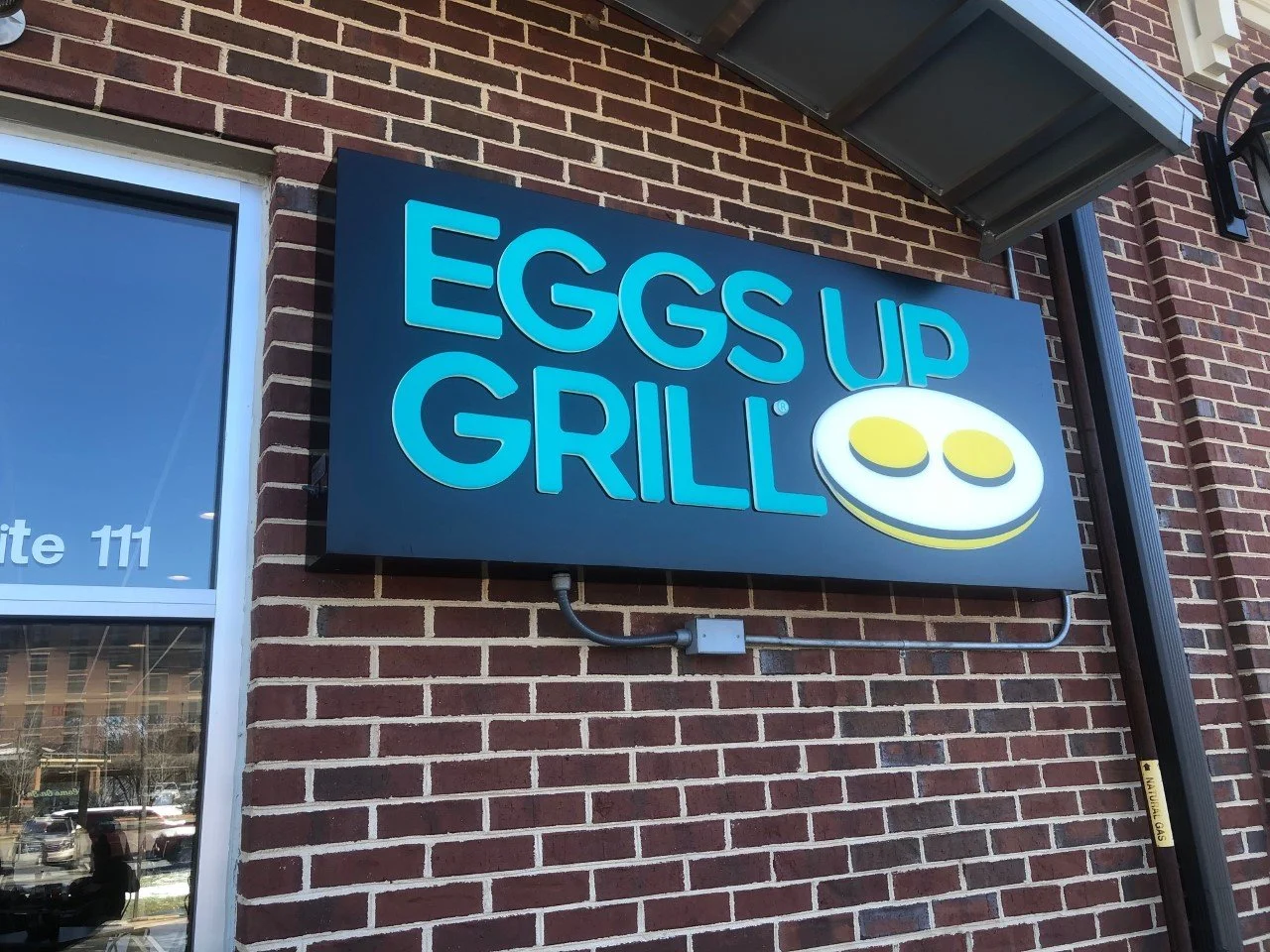 Sign for Eggs Up Grill with blue text and a smiling egg face logo on a brick wall.