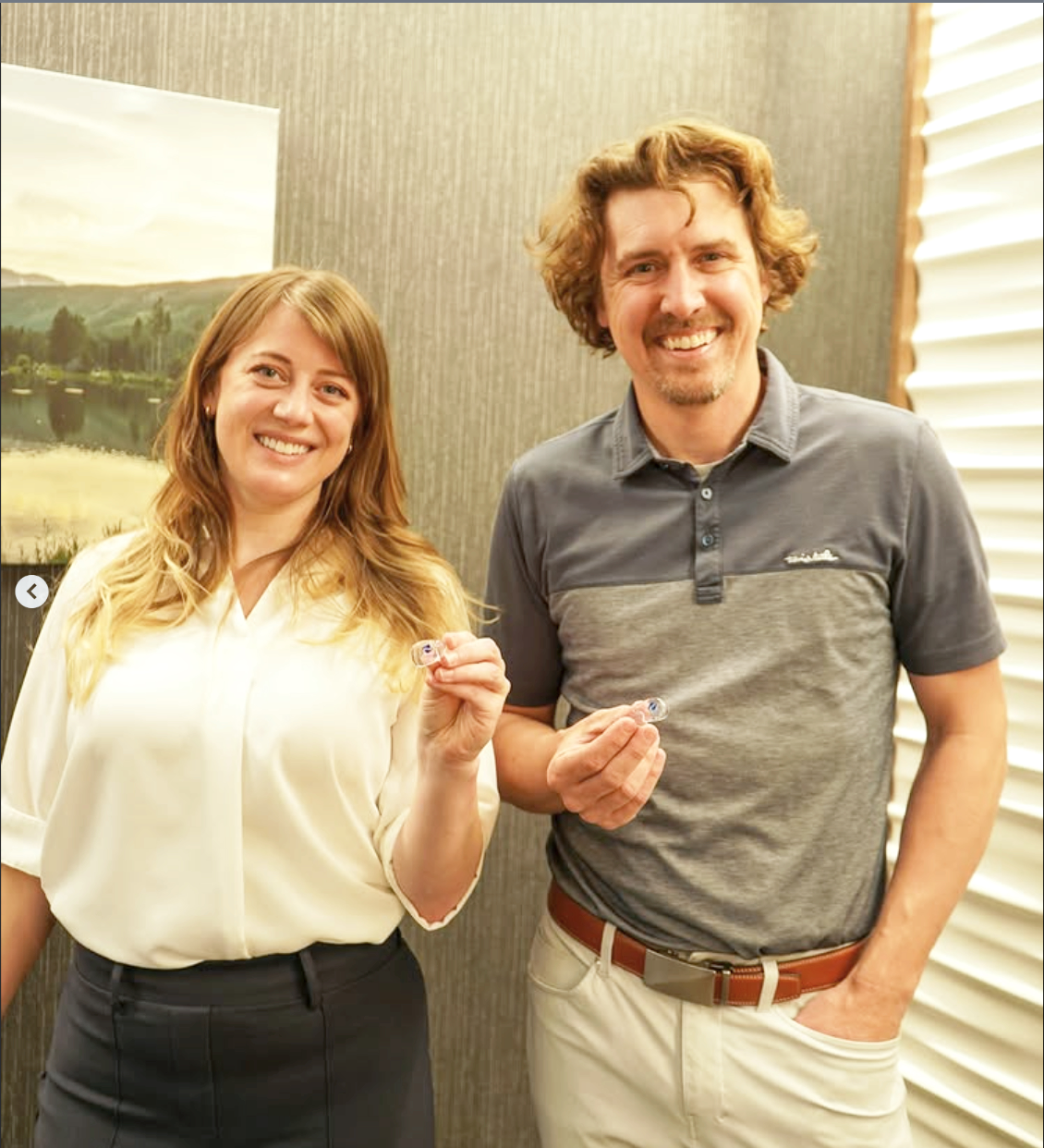 A woman and a man standing indoors, smiling, holding small objects, with a landscape picture on the wall behind them and window blinds on the right.