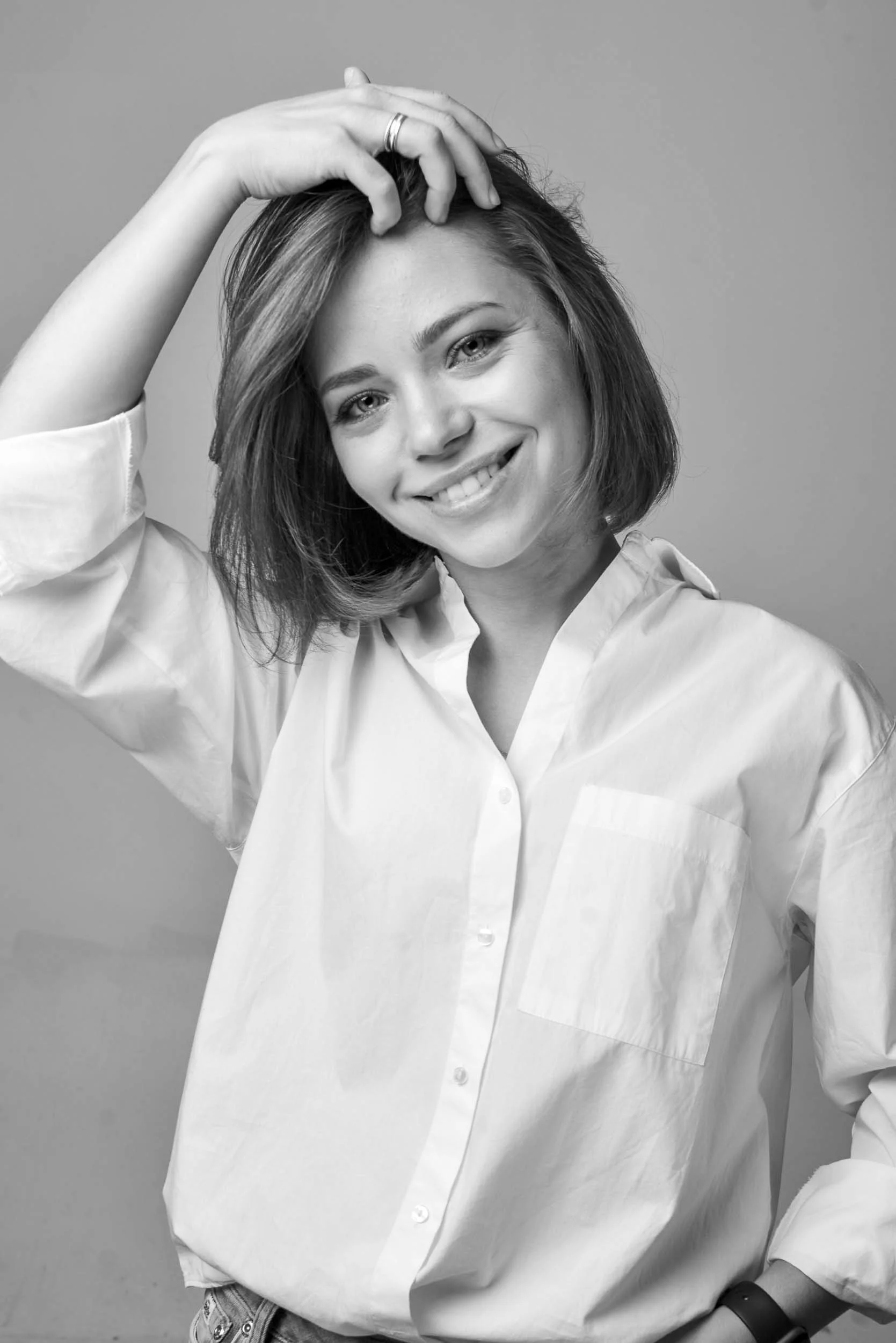 Black and white photo of a woman with short hair wearing a white button-up shirt, smiling, with her hand touching her hair.