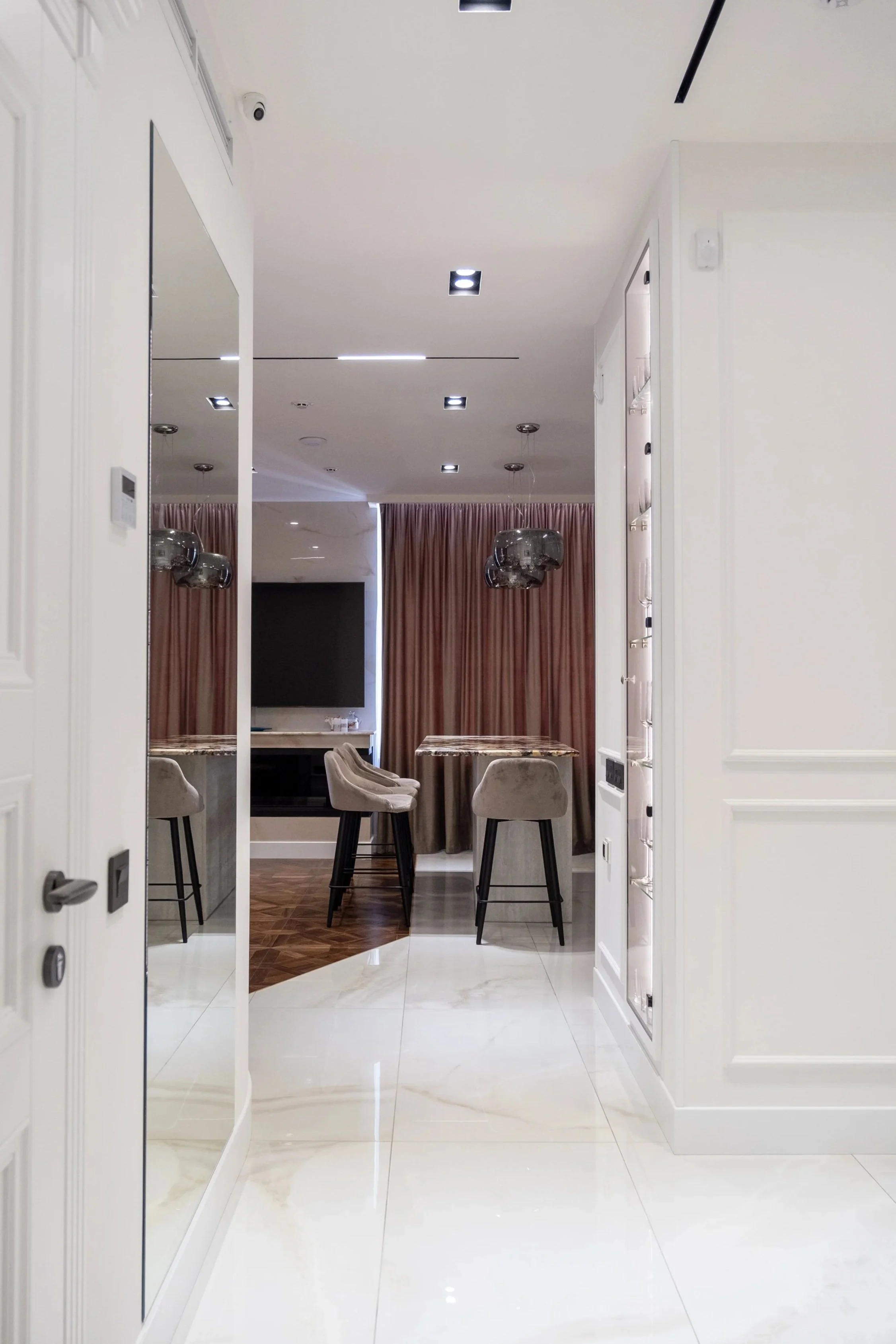 A modern, stylish indoor space with white walls, marble flooring, and a dining area with chairs and two tables. Pink curtains cover windows or a doorway in the background. The ceiling has recessed lighting and a security camera is visible in the upper corner.