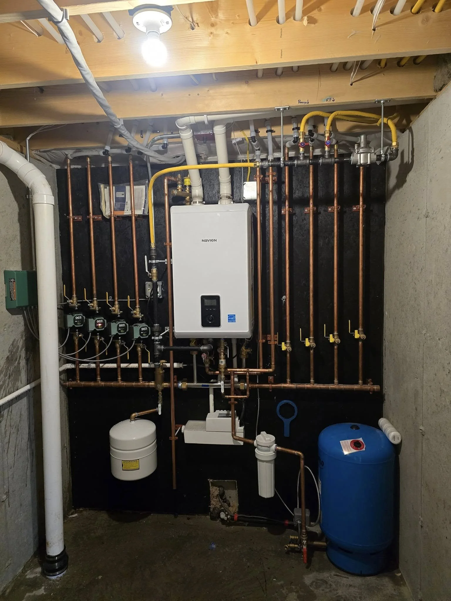 Image of a mechanical room showing plumbing, piping, and equipment. Features a Navien tankless water heater, copper pipes, a white water filter, a blue pressure tank, and various valves and fittings.