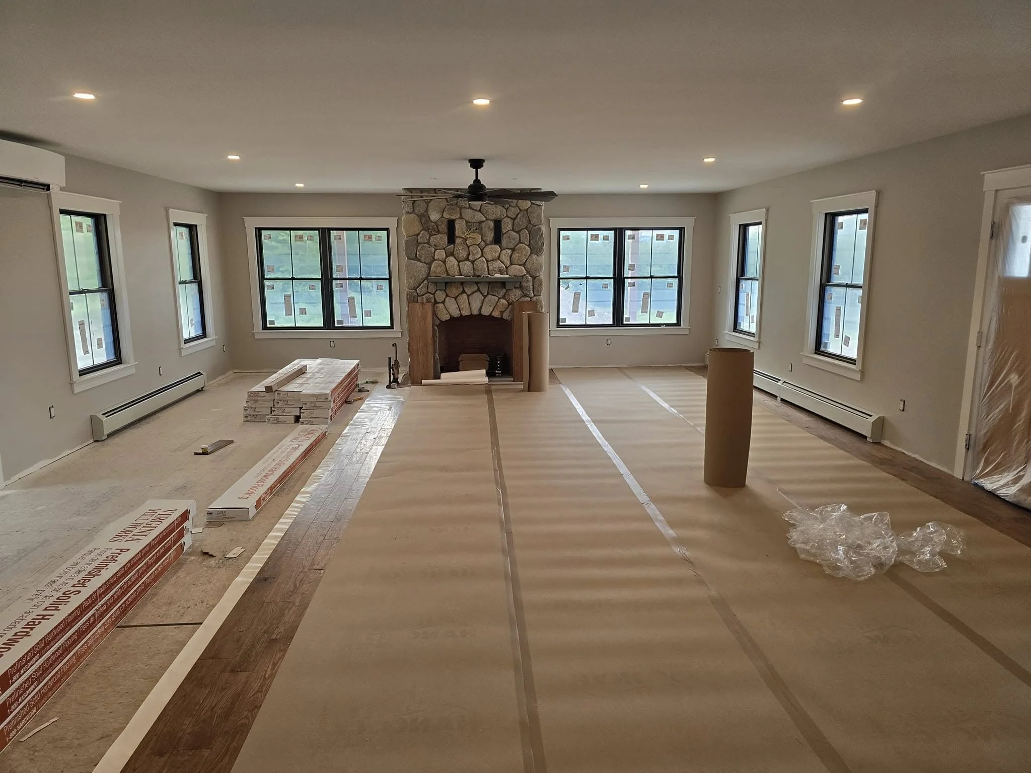 Living room under renovation with new hardwood flooring being installed, multiple windows, a stone fireplace, and construction materials on the floor.