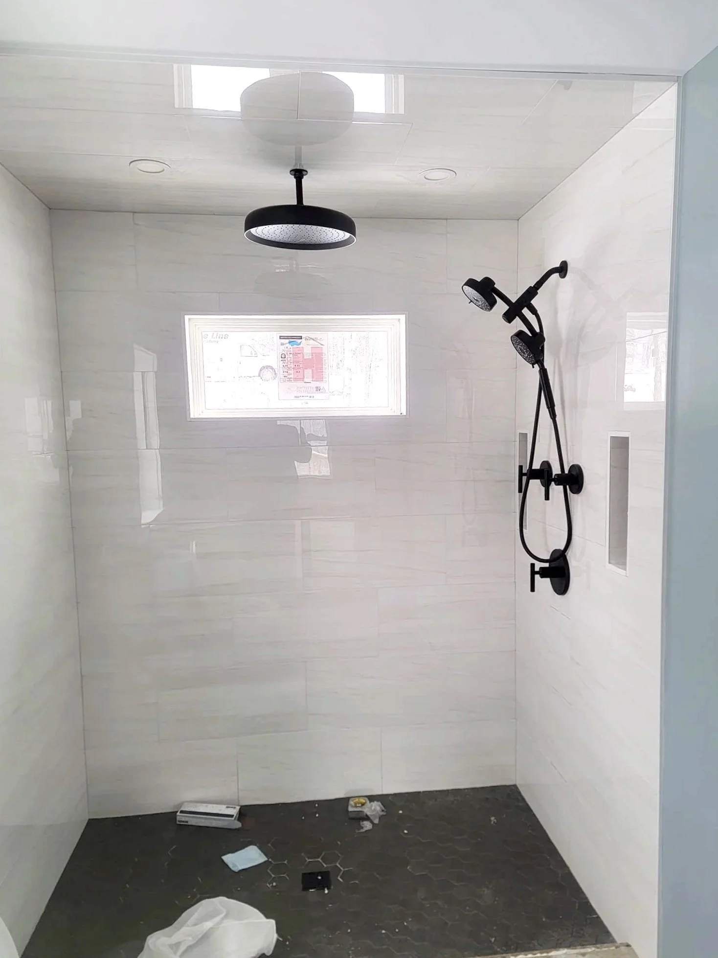 A partially finished bathroom shower area with white tiled walls, a black overhead shower, and a black showerhead with an adjustable arm. The floor is covered with hexagonal black tiles, and there are some debris and items scattered on the floor, including a box, a crumpled paper towel, and some small objects. A window is visible on the back wall.