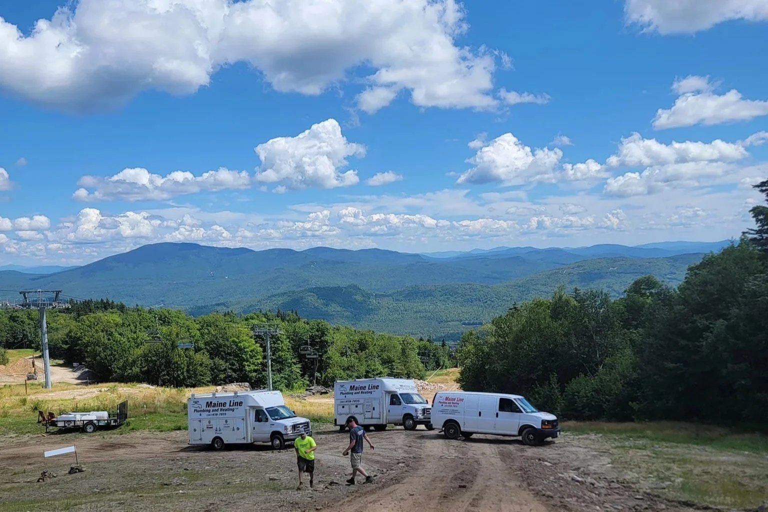 Three service vans from Maine Line Plumbing & Heating parked on a dirt area with two people standing and talking in front of the vans; a scenic backdrop of green forested mountains and a blue sky with scattered clouds.