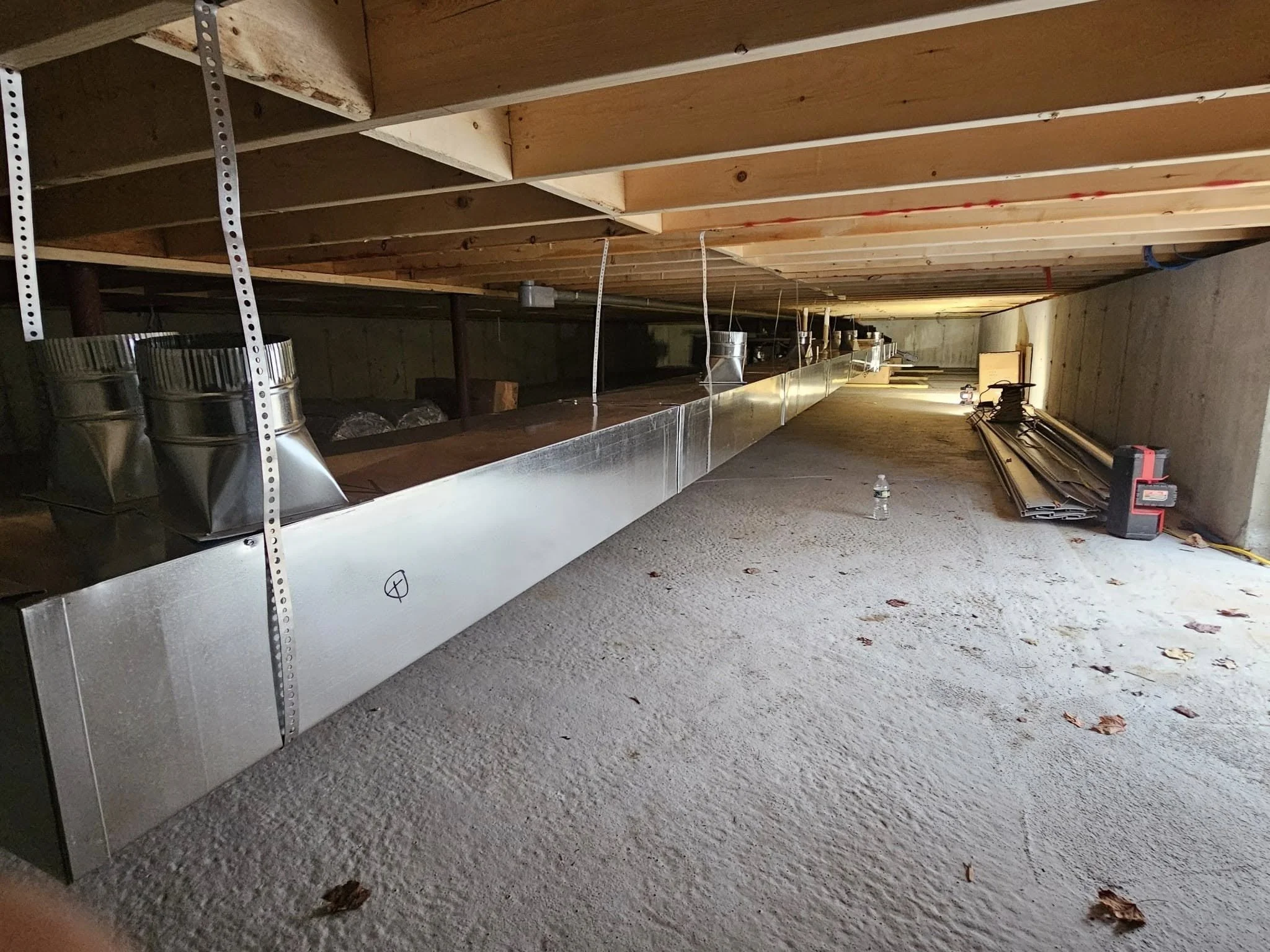 Construction site in a basement with exposed wooden ceiling beams, metal ductwork, and miscellaneous construction tools and materials scattered across the unfinished floor.