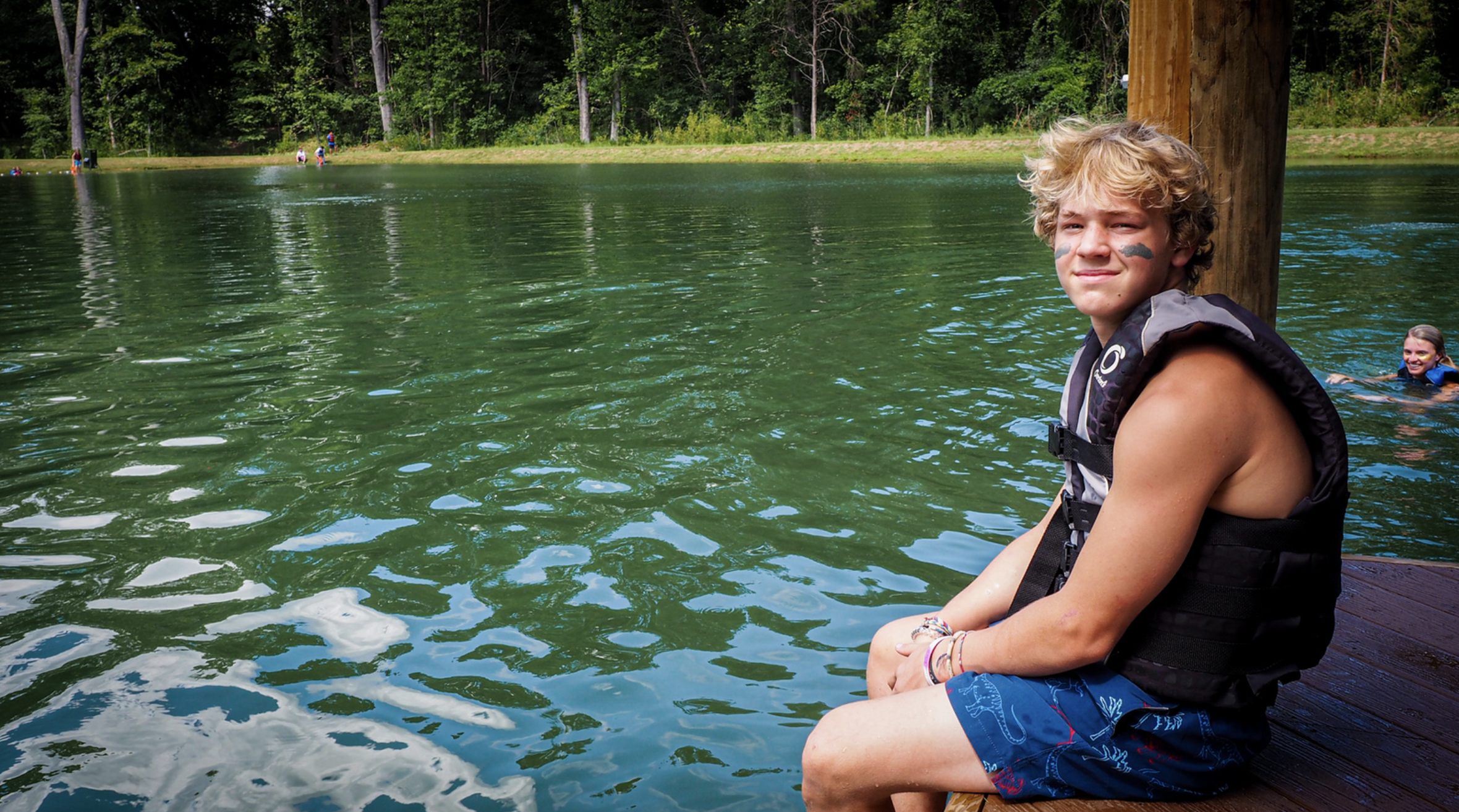 A boy sits on a dock by a lake with a life vest on, looking at the camera with a slight smile, while a girl swims in the background at Riverstone at Canaan.