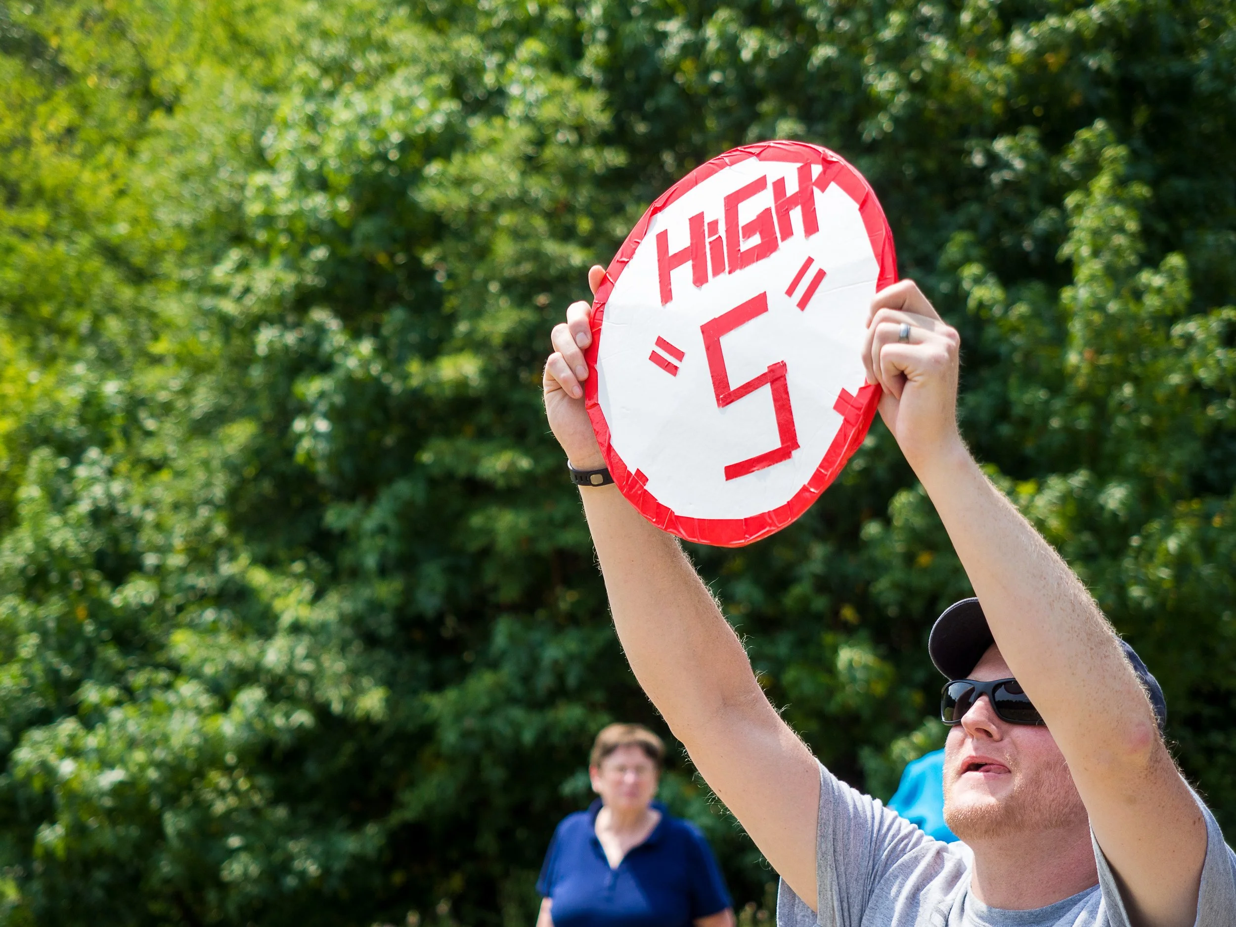 A person holding a red and white speed limit sign that reads 'HIGH 5' outdoors, with other people in the background and surrounded by green trees at Riverstone at Canaan.