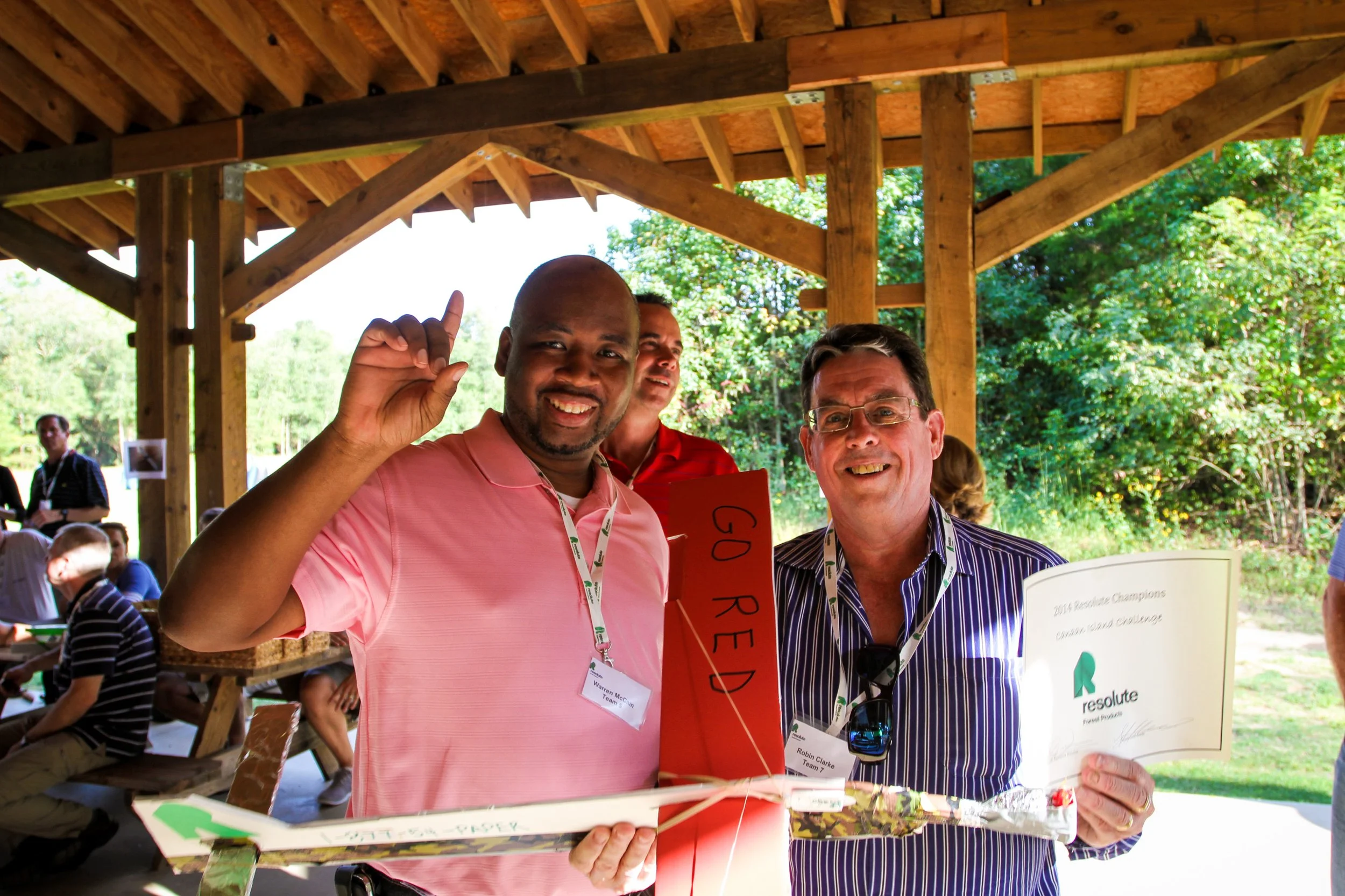 Two men smiling and holding certificates at an outdoor event under a wooden pavilion, with other people sitting and standing nearby, greenery in the background at Riverstone at Canaan.