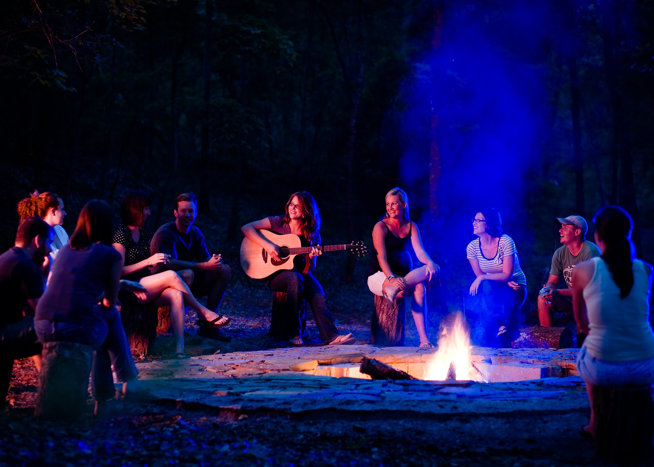 Group of people sitting around a campfire in a forest at night, with some playing guitar and smiling at Riverstone at Canaan.