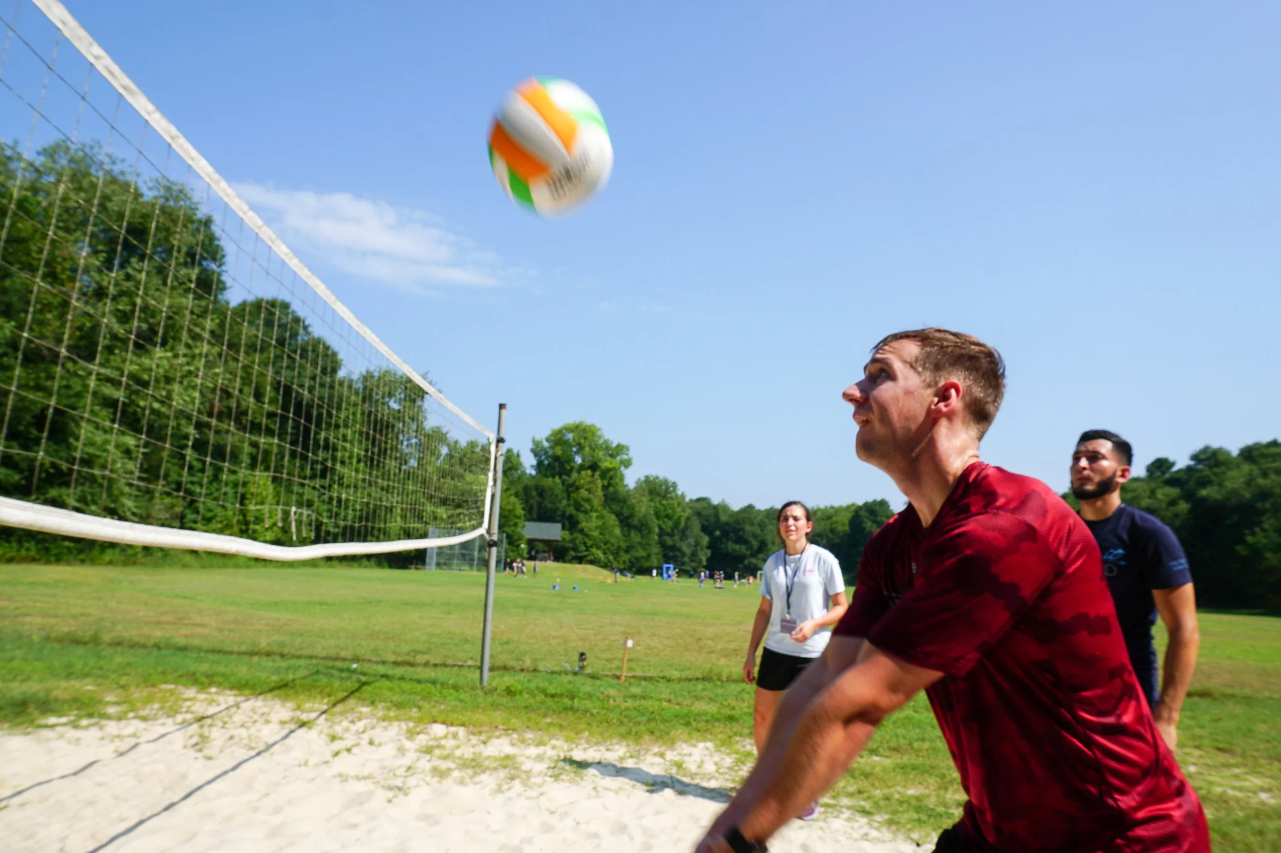A man in a red shirt hits a volleyball over a net during an outdoor game on a sunny day, with other people watching in the background at Riverstone at Canaan.