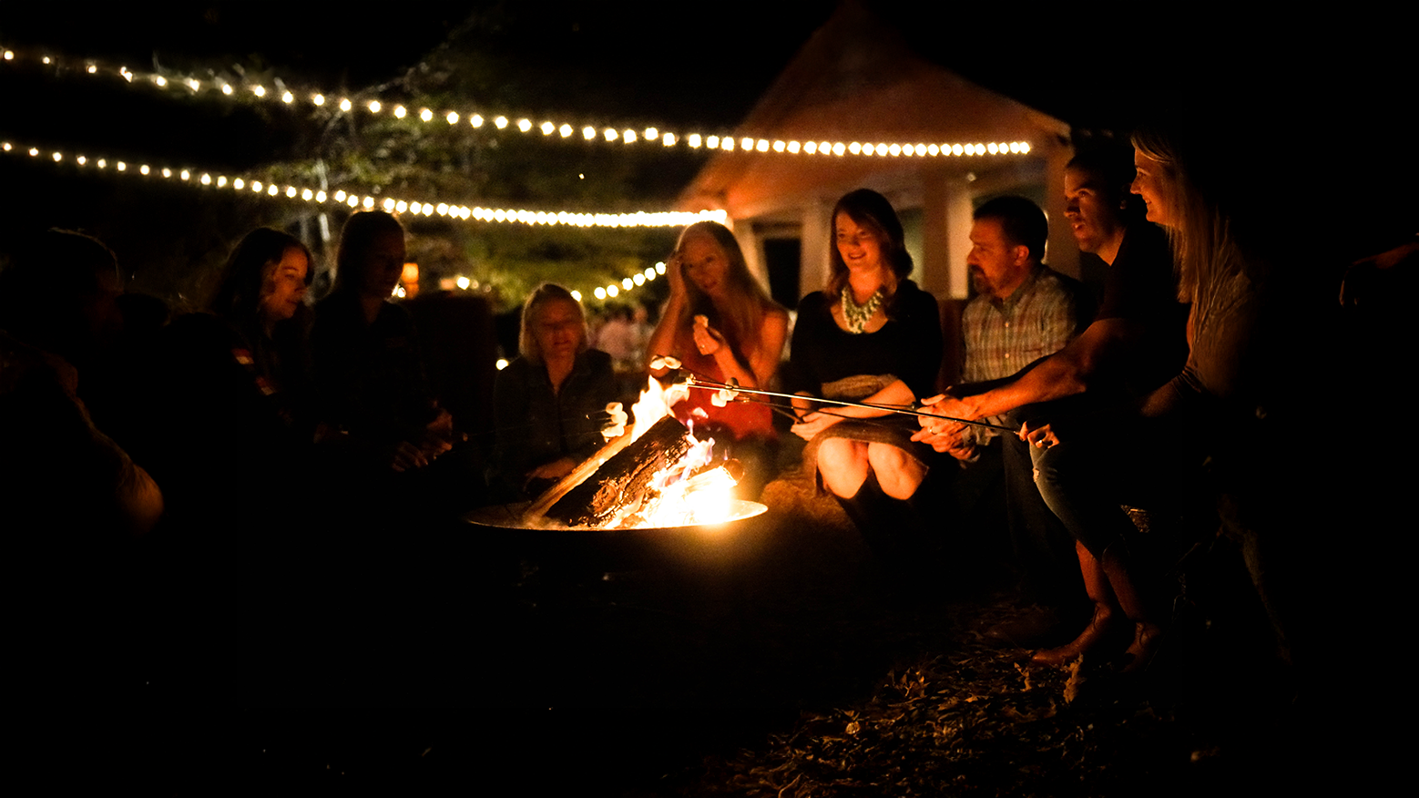 Group of people around a campfire at night with string lights overhead, smiling and roasting marshmallows at Riverstone at Canaan.