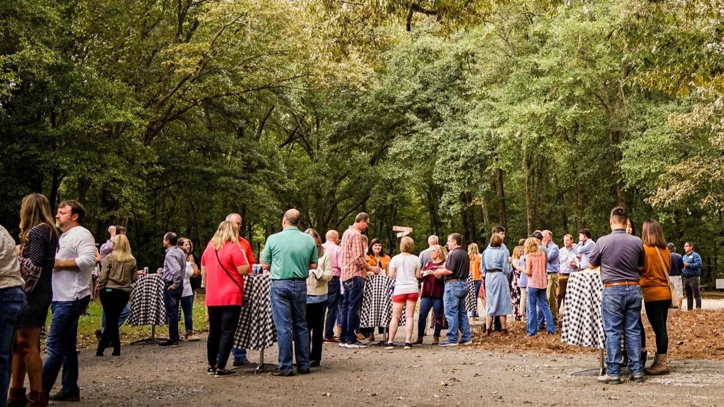 People gathered outdoors in a wooded area with picnic tables covered in checkered tablecloths, engaged in conversation at a social event at Riverstone at Canaan.