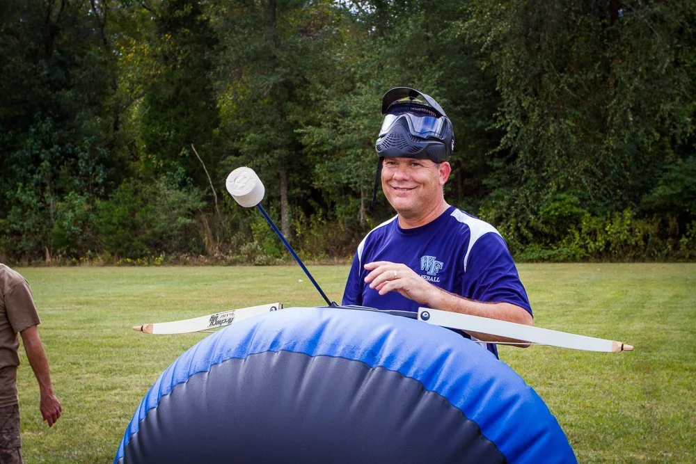 A man wearing a blue sports shirt and safety goggles smiles while standing outdoors near trees, holding a model airplane with a parachute attached to the back at Riverstone at Canaan.
