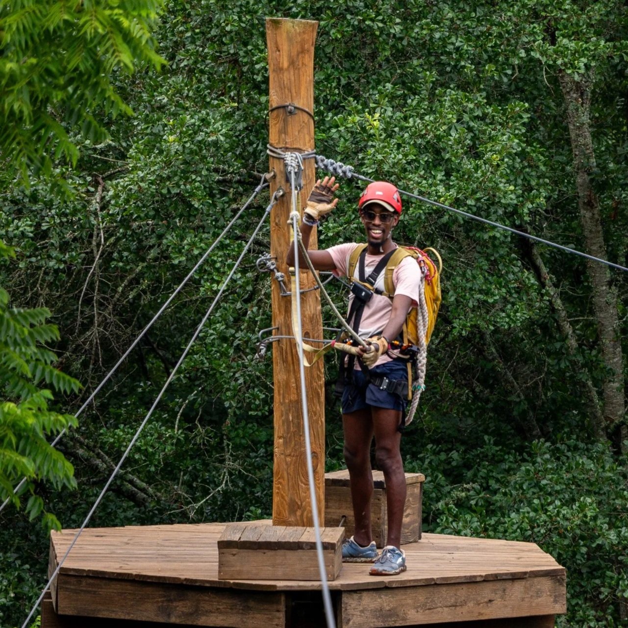 A man standing on a wooden platform at a treetop adventure course, holding onto safety ropes, wearing a helmet, sunglasses, gloves, a backpack, and adventure harness, surrounded by dense green forest at Riverstone at Canaan.