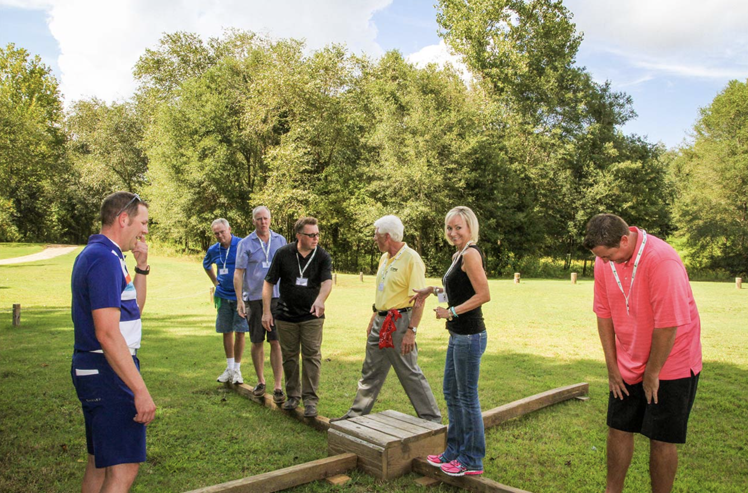 Group of people outdoors on a sunny day, engaging in a team-building activity involving walking on wooden planks placed closely together on the grass at Riverstone at Canaan.