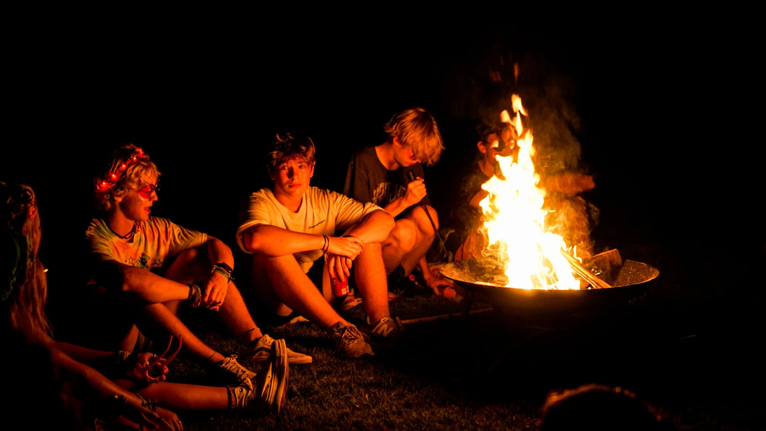 Group of young people sitting around a campfire at night, some with drinks, enjoying each other's company at Riverstone at Canaan.