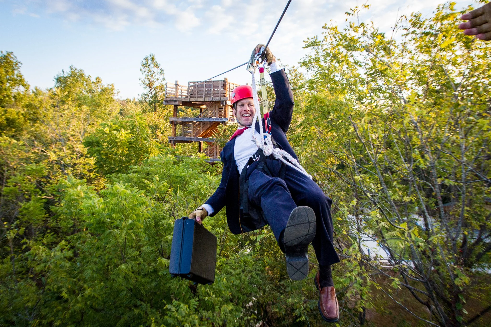 A man wearing a red helmet and harness ziplining through a green forest, holding onto the zipline handle with one hand and smiling at Riverstone at Canaan.