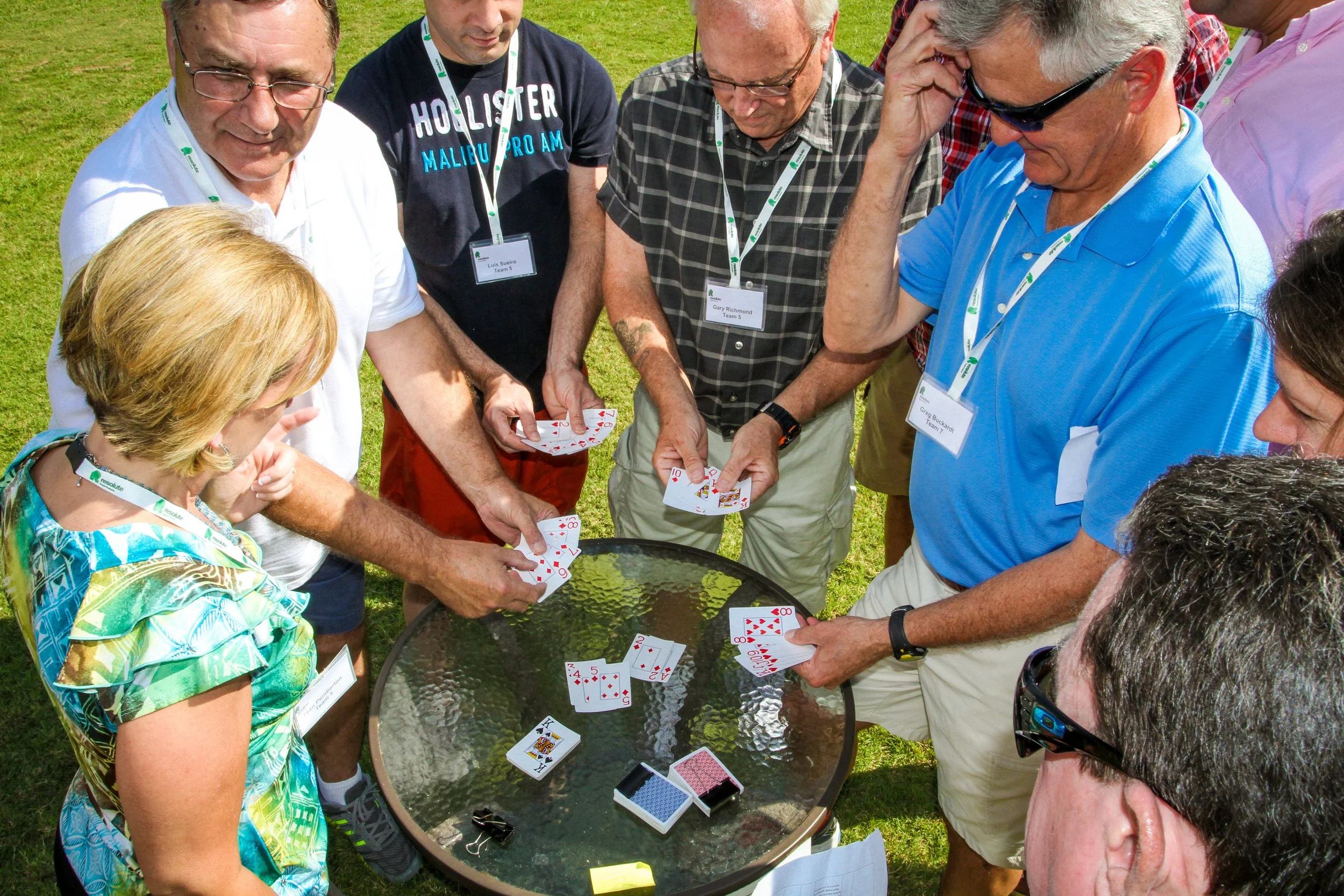A group of people playing cards outdoors around a glass table in a grassy area, wearing lanyards with name tags at Riverstone at Canaan.