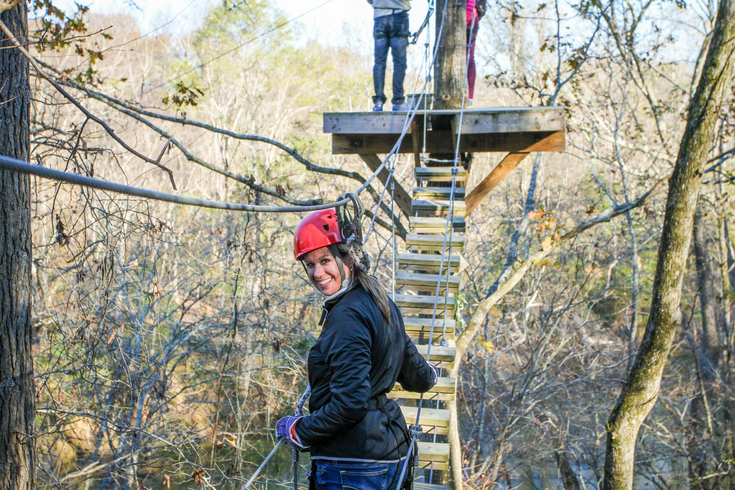 A woman with a red helmet smiling while crossing a suspension bridge in the woods with two other people ahead on the bridge at Riverstone at Canaan.
