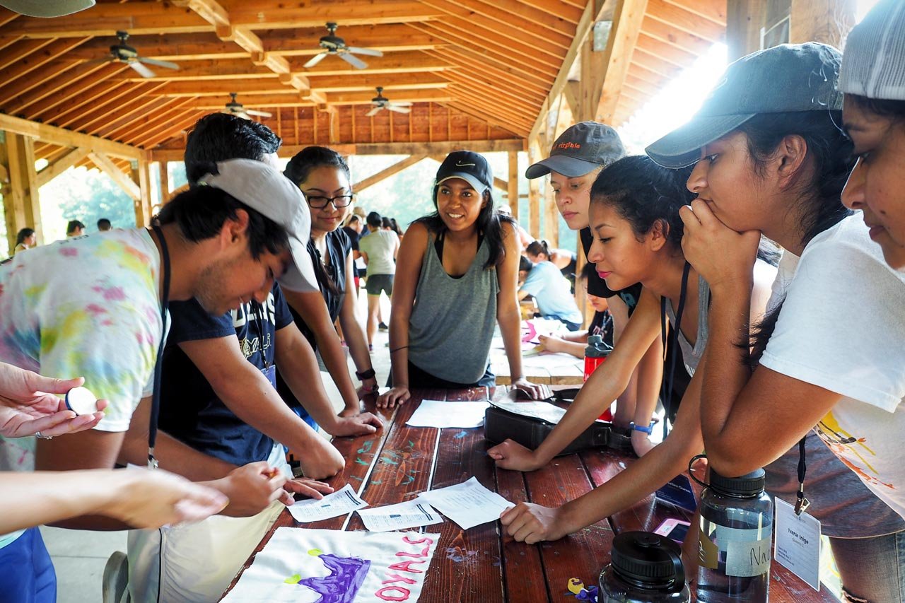 Group of young people gathered around a table under a wooden pavilion, looking at papers and engaged in a discussion or activity at Riverstone at Canaan.