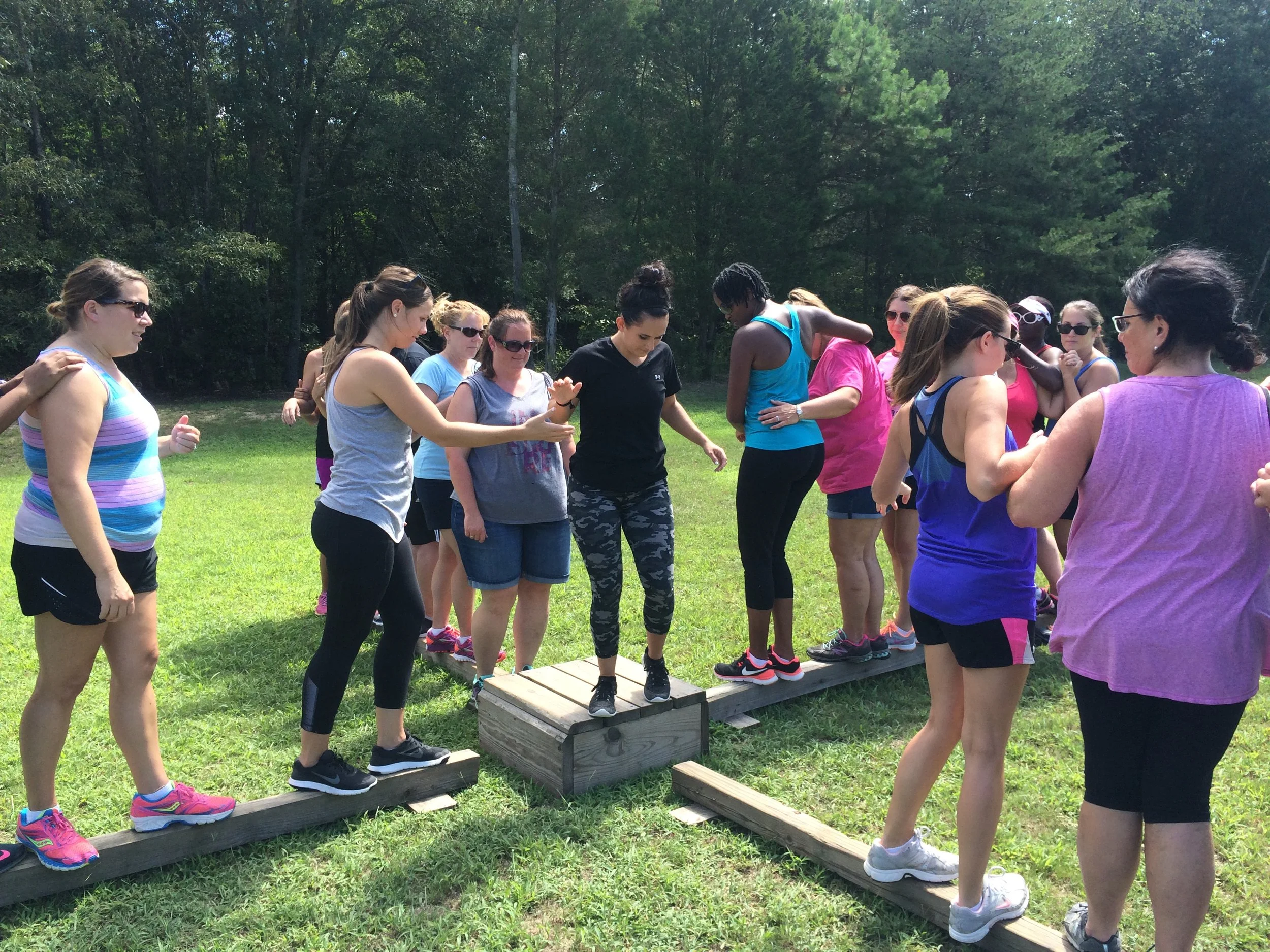 A group of women participating in an outdoor team-building activity crossing a wooden balance beam in a grassy park with trees in the background at Riverstone at Canaan.
