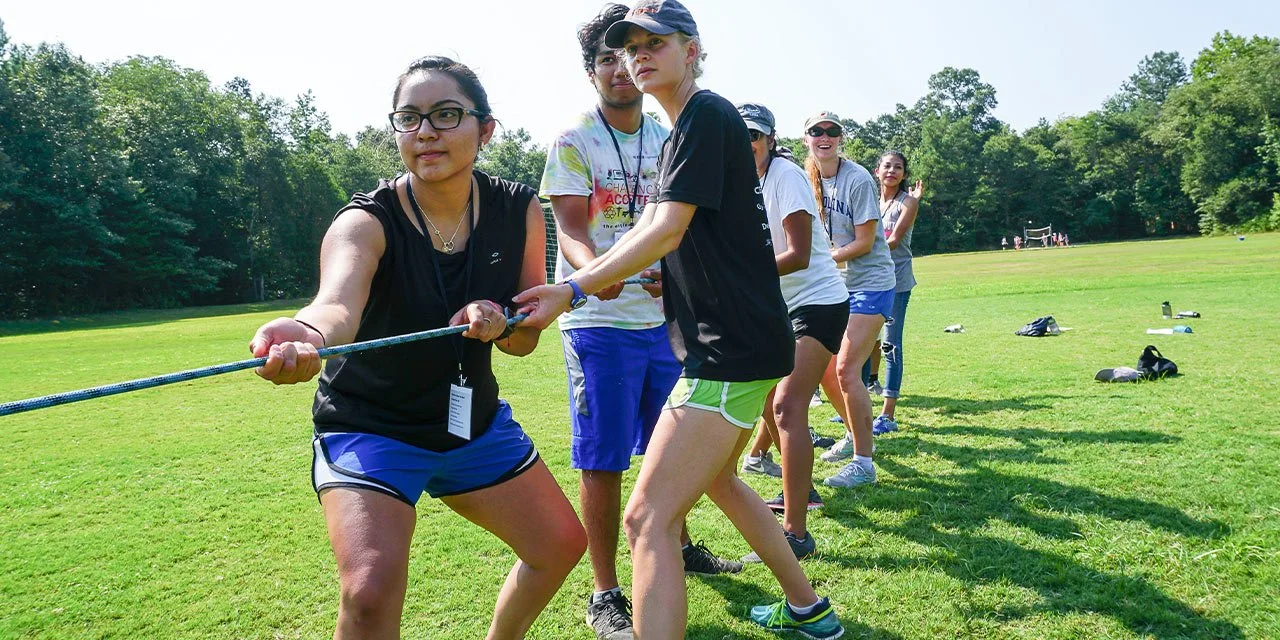 Group of young women participating in a tug-of-war game outdoors on a grassy field, with trees in the background at Riverstone at Canaan.