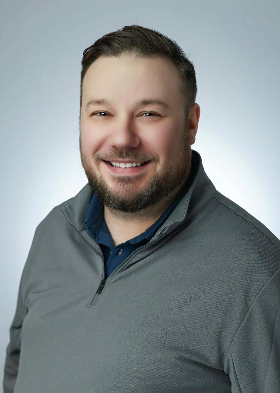 A smiling man with short dark hair and a beard, wearing a gray quarter-zip jacket over a dark blue shirt, posing against a plain light gray background.