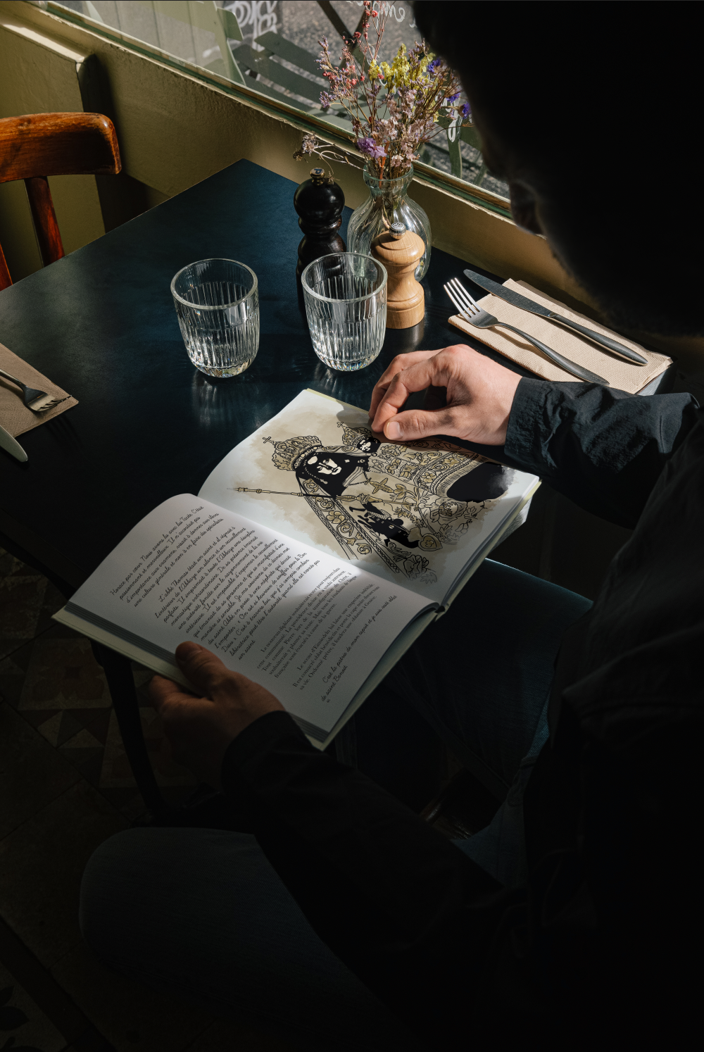 Une personne lit un livre illustré à une table de restaurant près d'une fenêtre, avec des verres d'eau, des assiettes, une bouteille de sel, une poivrière, un moulin à poivre, et un vase avec des fleurs séchées.