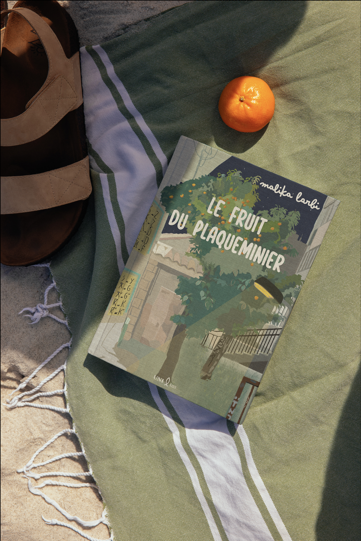 Livre intitulé 'Le Fruit du Plaque Miner' posé sur une serviette à carreaux vert et blanc, avec une mandarine, un sac en cuir brun, et un tissu à franges blanches. La scène est éclairée par la lumière du soleil.