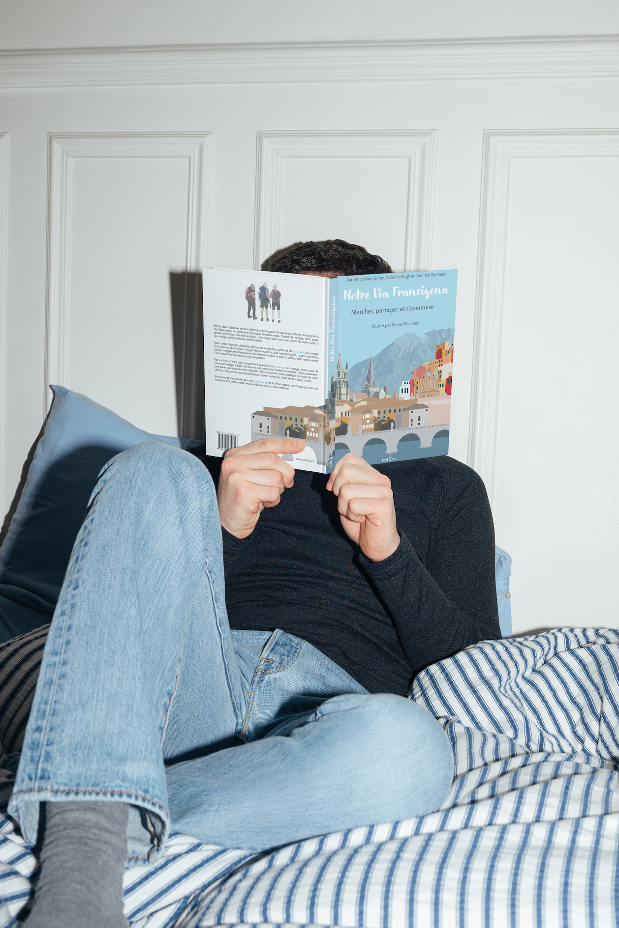 Personne allongée sur un lit, tenant un livre intitulé 'Notre Via Francigena' devant le visage, avec un mur blanc en arrière-plan.