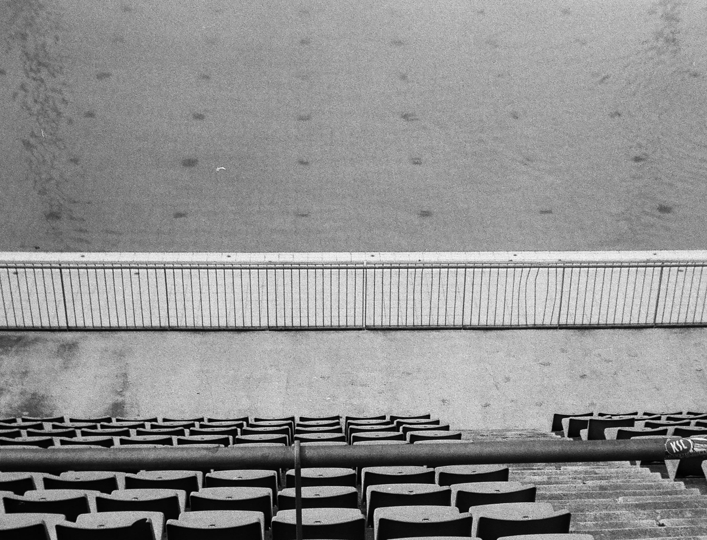 Vue d'une enceinte de stade vide avec plusieurs rangées de sièges en plastique et un terrain au centre, vue en noir et blanc.