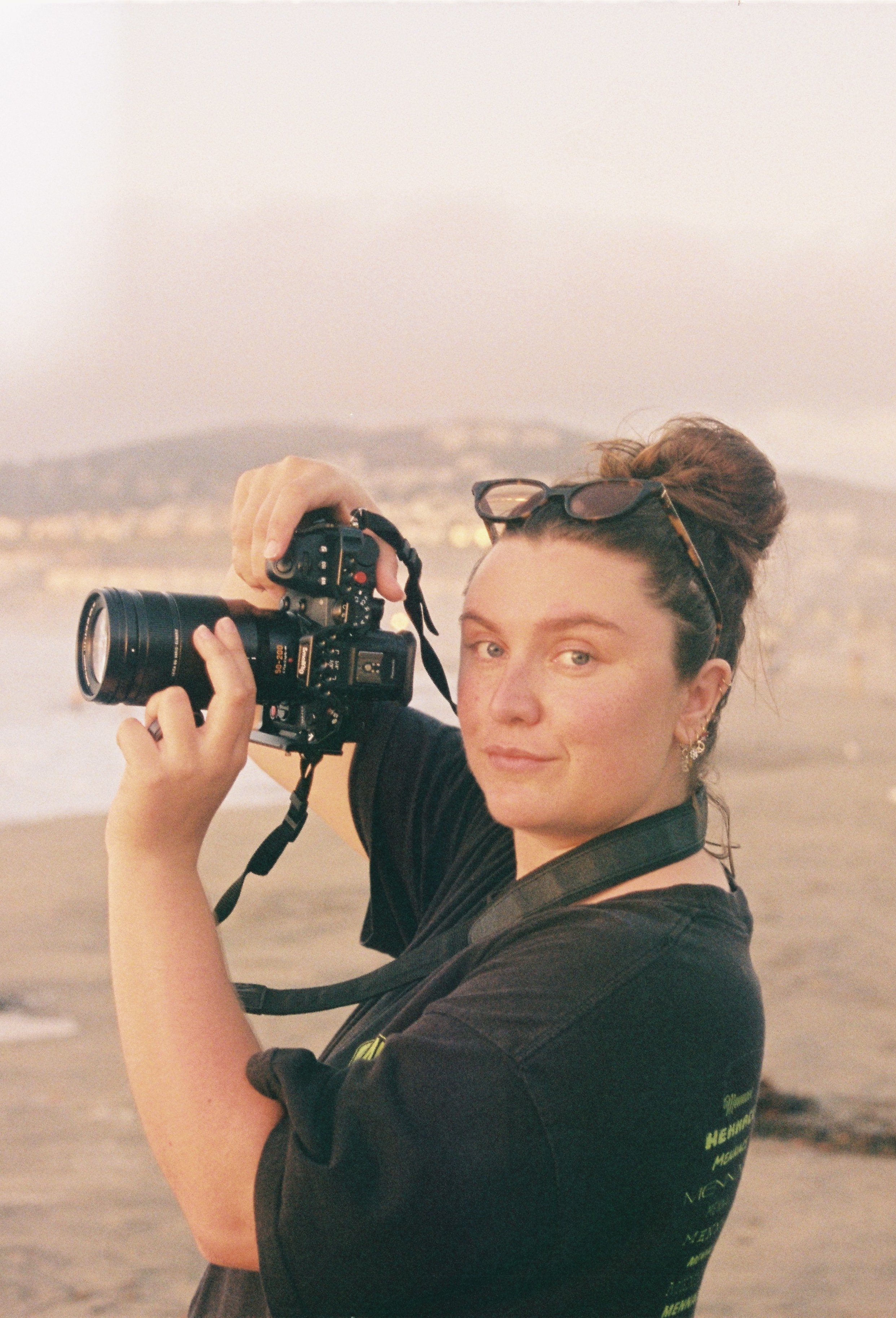 Une femme avec des cheveux attachés en chignon portant des lunettes sur sa tête, tenant un appareil photo numérique lors d'une sortie à la plage.