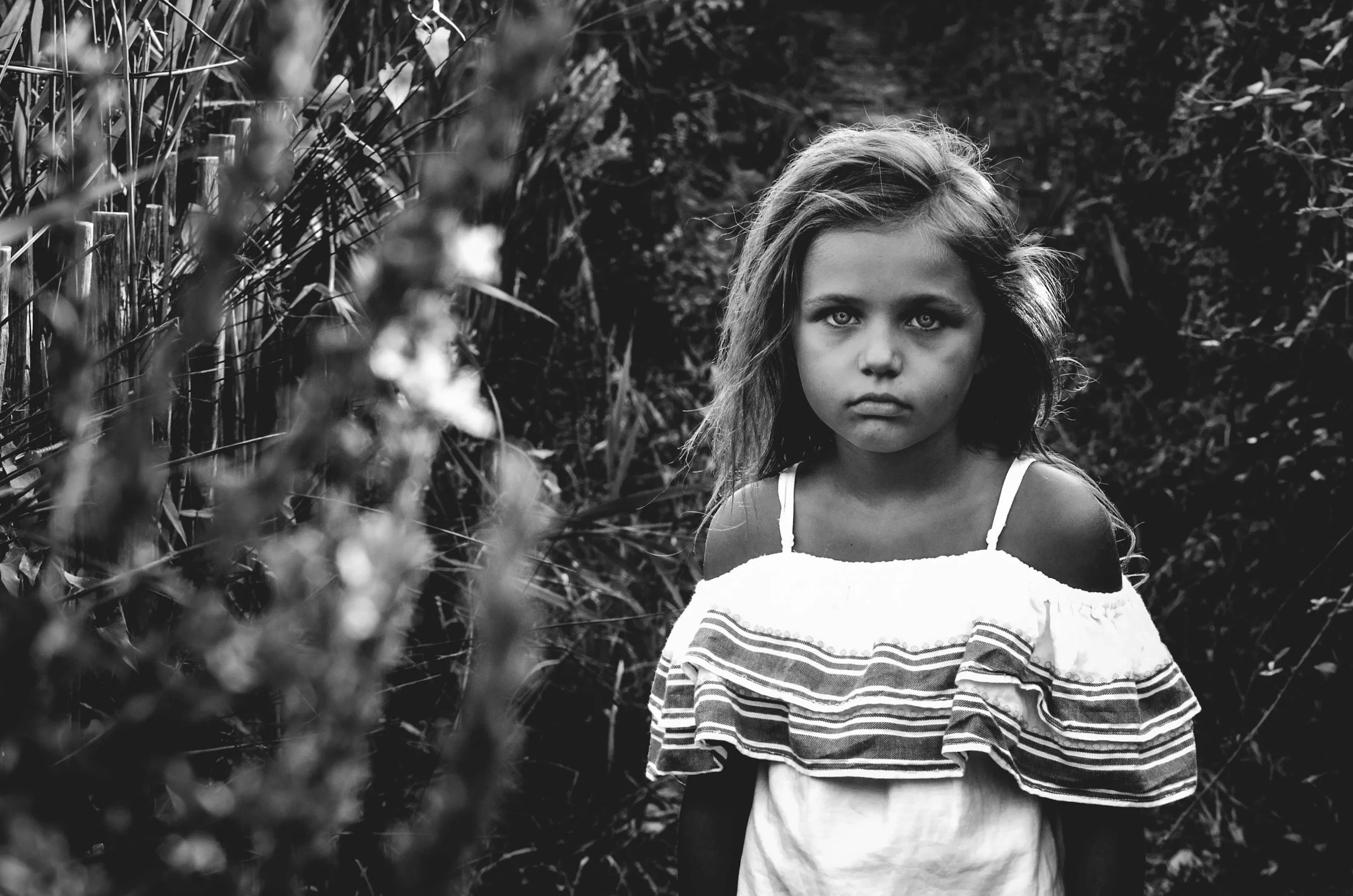 A young girl with long hair and a serious expression stands outdoors in a natural setting among bushes and foliage, wearing a striped shirt.