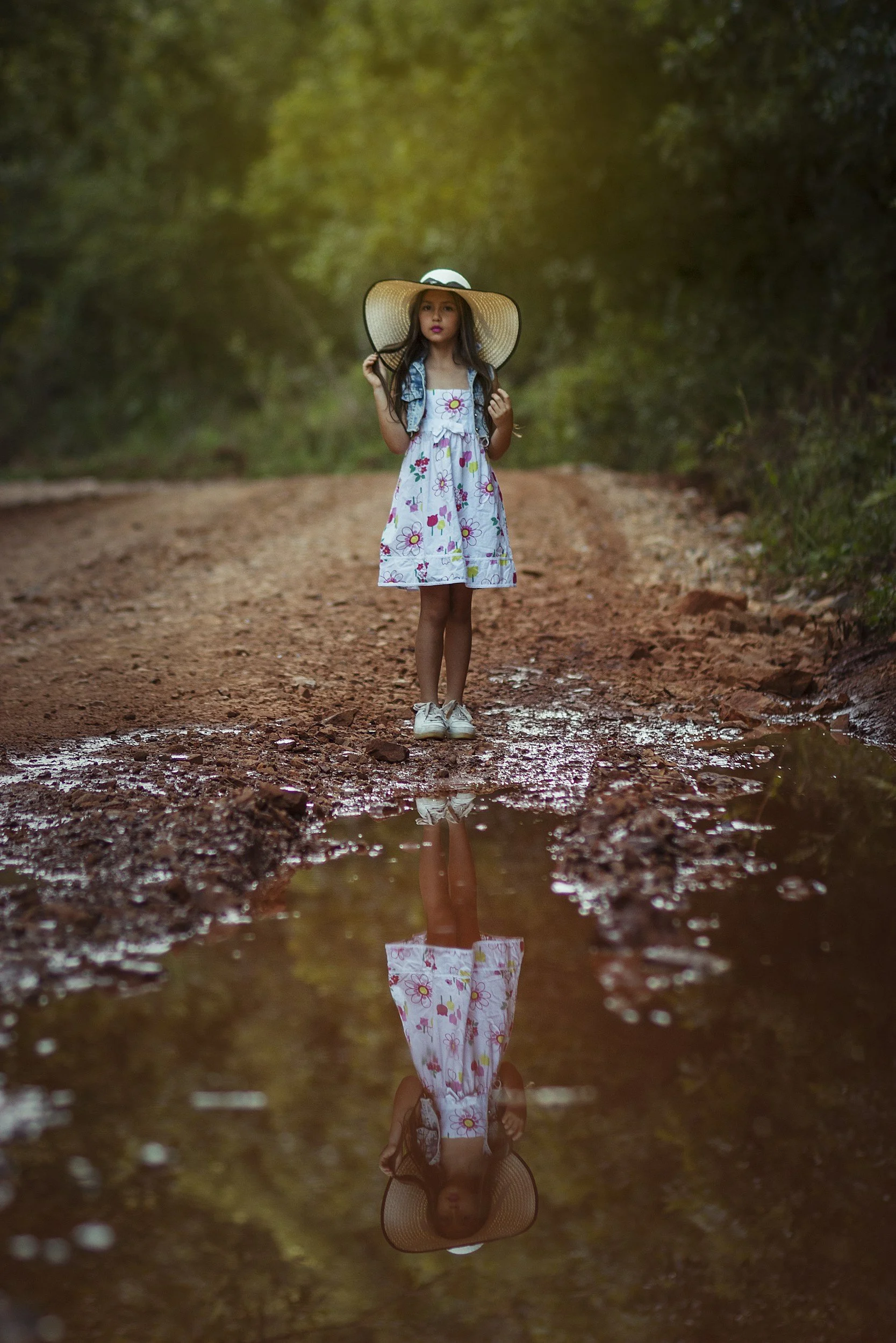 Young girl wearing a floral dress and wide-brimmed hat standing outdoors on a dirt path, with her reflection visible in a puddle on the ground.