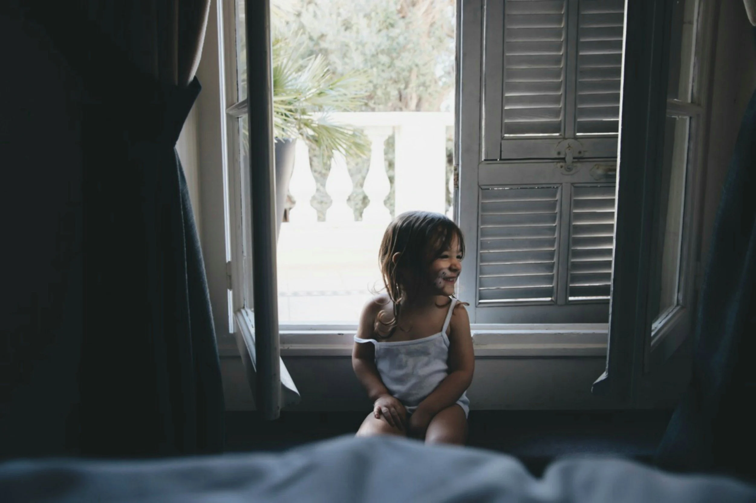 A young girl with long, brown hair sitting by an open window with wooden shutters, smiling inside a house.
